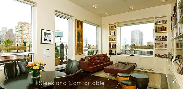 Modern living room with floor-to-ceiling windows, burgundy sofa, built-in shelving, and city skyline views, designed by Marjorie Hilton.