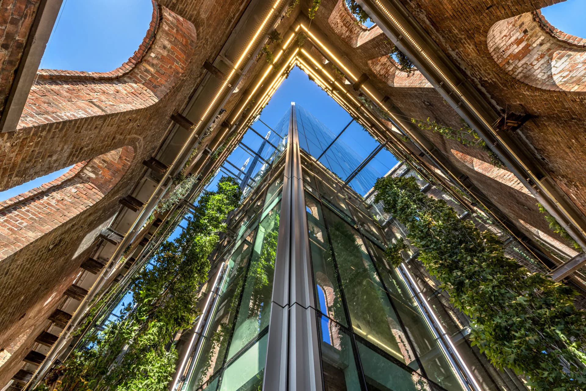 Atrium with soaring glass and steel structure framed by brick walls, lush climbing ivy, and clear blue sky visible above, by Mark Faulkner.