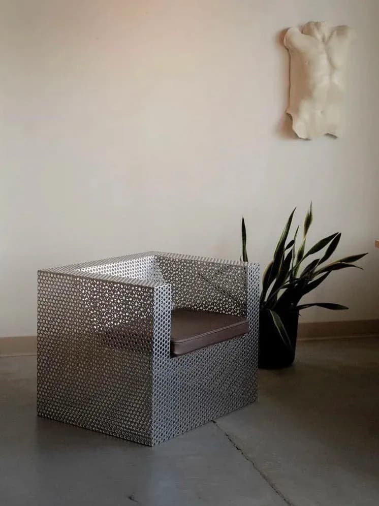 Modern sculptural chair with perforated metal frame and dark seat cushion, styled with black sculptural plant and white wall art by Mark Malecki.