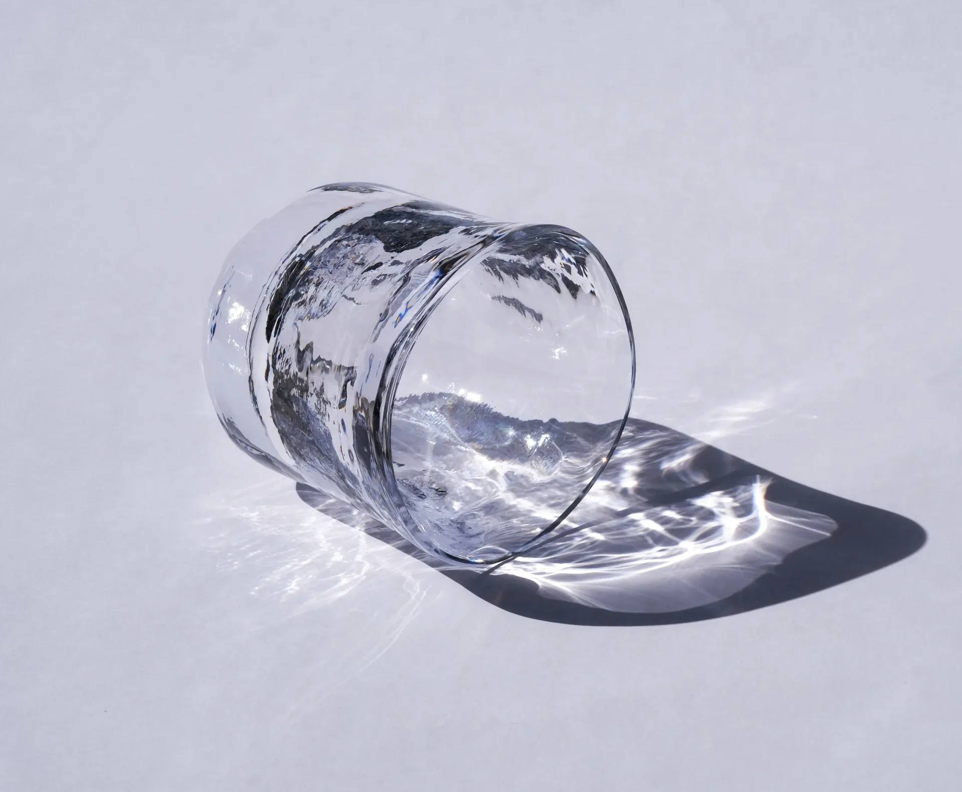 A transparent glass cylinder tilted on its side with water spilling out, creating dramatic light reflections and shadows against a pale background by Mark Pavlovits.