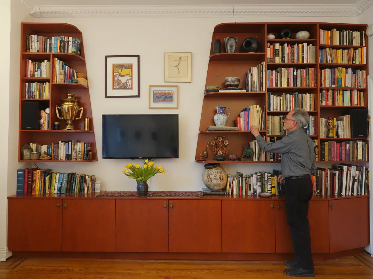 Custom built-in cabinetry with wood shelving, integrated TV, and storage in warm cognac tones flanking framed artwork and decorative objects by Mark Rosenhaus.