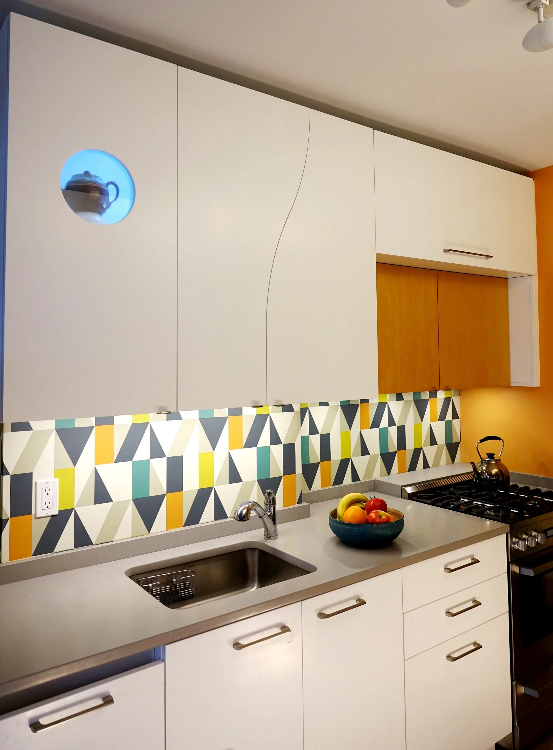 Modern kitchen with white cabinetry, geometric patterned backsplash in yellow, teal, and charcoal, stainless steel appliances, and warm orange accent wall by Mark Rosenhaus.