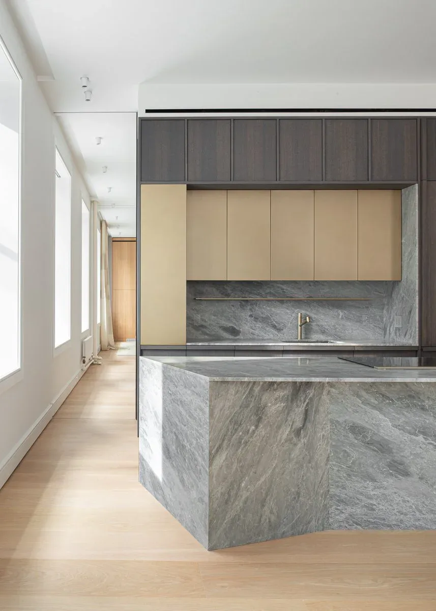Contemporary kitchen with curved marble island, cream cabinetry, dark upper cabinets, and light wood flooring in a minimalist design by Marlene Bednarz.