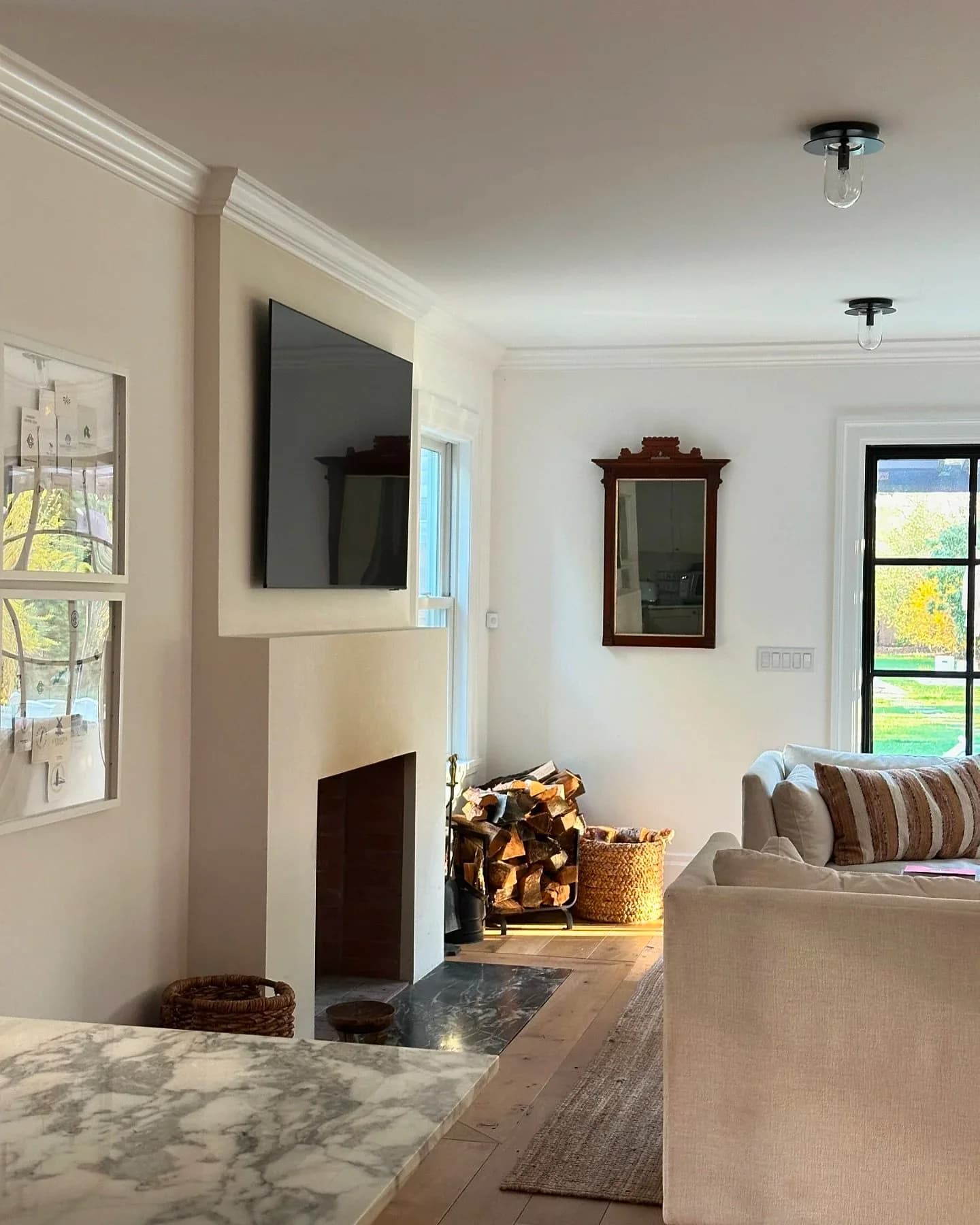 Bright, airy living room with marble countertop, fireplace with dark surround, wall-mounted TV, cream sofa, and large windows overlooking landscape by Martina Montalbetti.