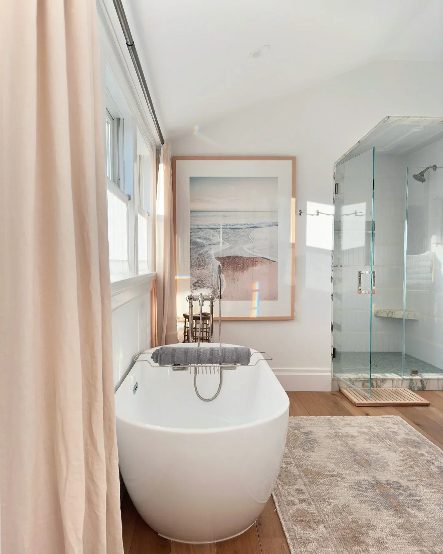 Modern spa bathroom featuring a white freestanding soaking tub, glass-enclosed shower, soft pink curtains, and serene coastal artwork, designed by Martina Montalbetti.
