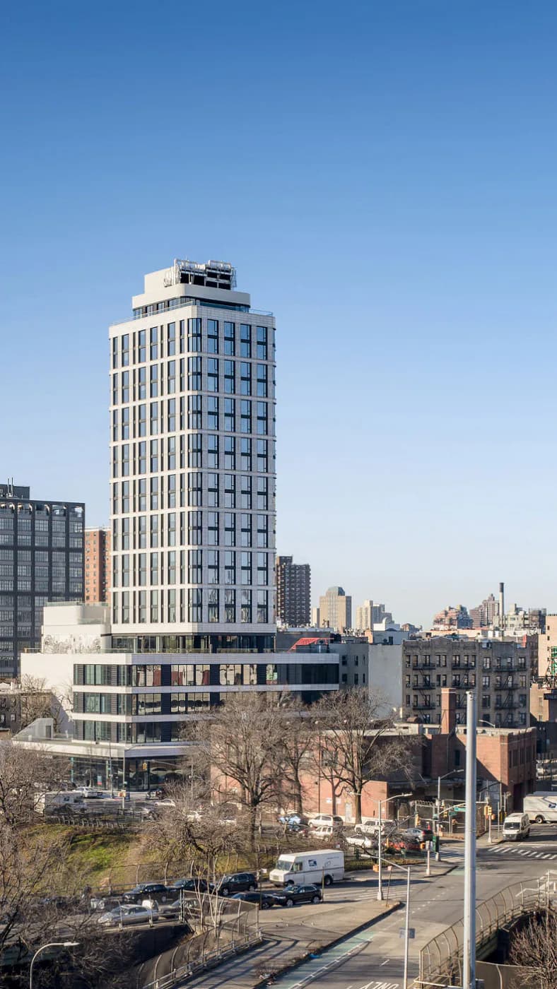 Contemporary high-rise residential tower with vertical ribbed facade, blue-toned glass windows, and integrated ground-level retail, set against Manhattan skyline by Matilde Pietrabissa.