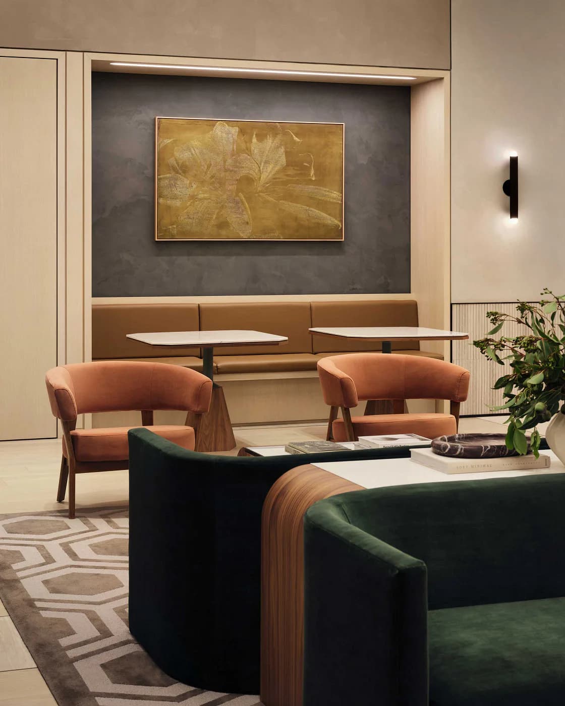 Contemporary lounge featuring terracotta upholstered curved chairs, dark green seating, geometric patterned rug, and built-in bench seating beneath gold-toned artwork by Matilde Pietrabissa.