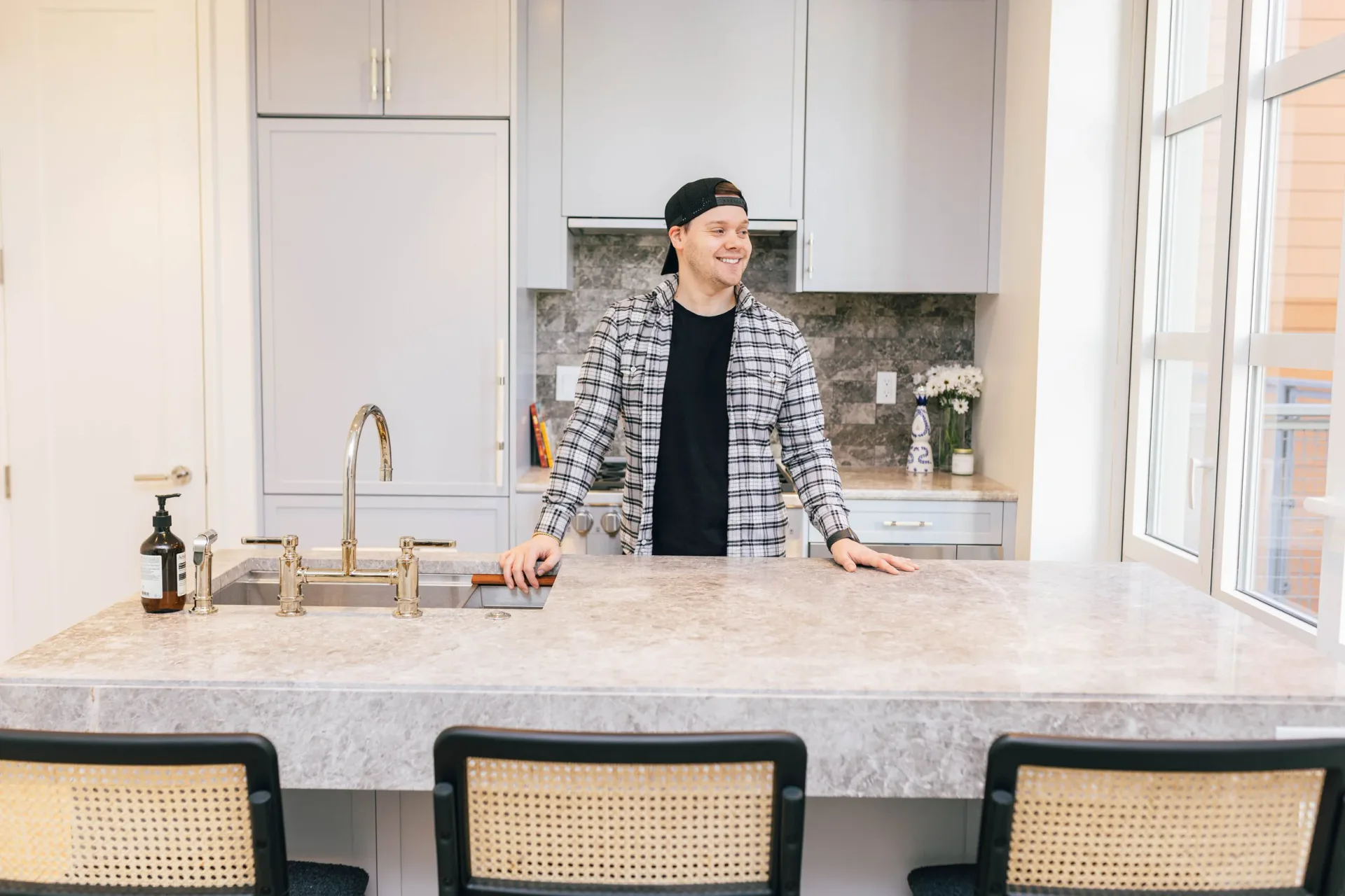 Modern kitchen with white cabinetry, marble waterfall countertop, and woven rattan bar stools by Matt Labombard.