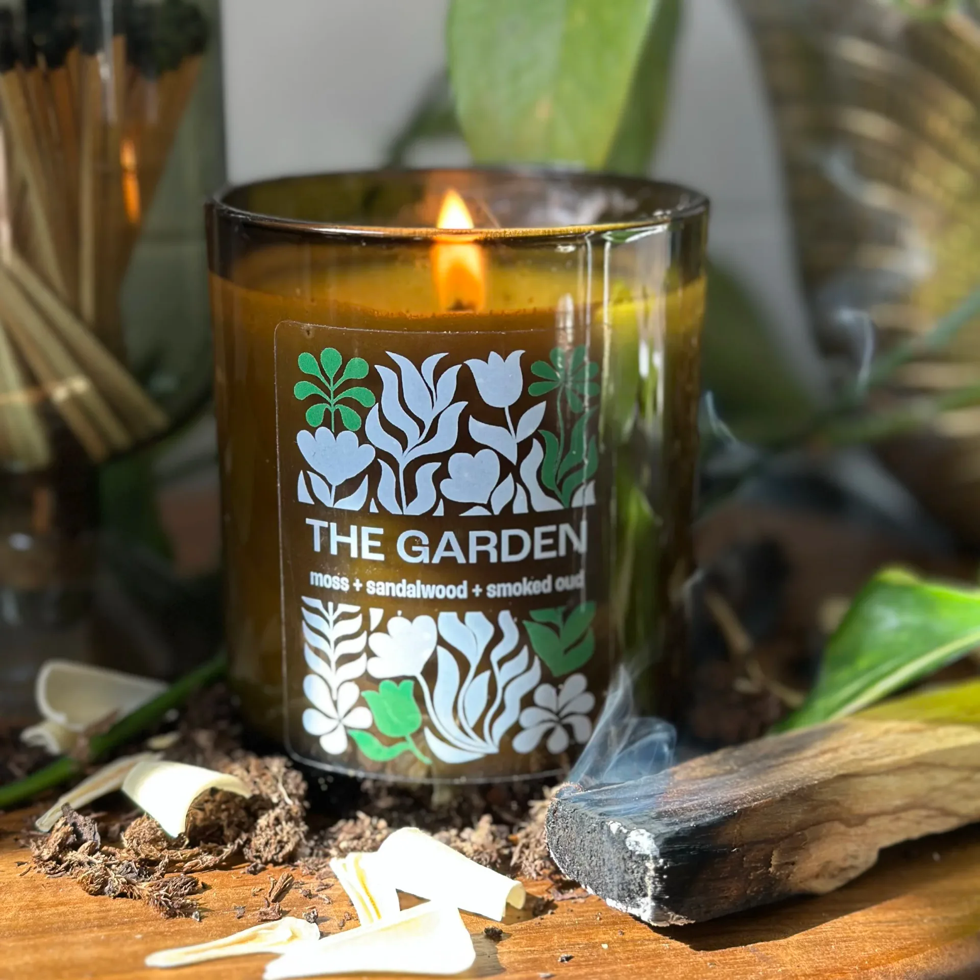 Dark glass candle jar with botanical label design featuring white floral illustrations and green accents, styled on wooden surface with natural elements by Matt Labombard.