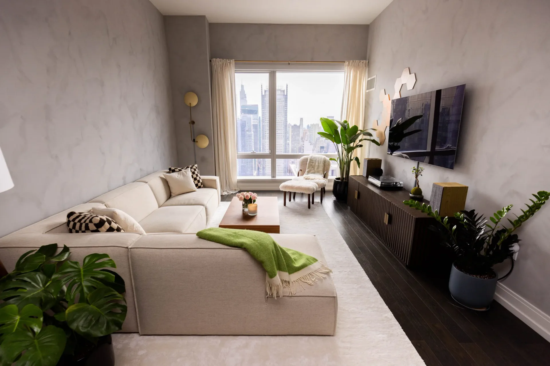 Modern living room with cream sectional, dark media console, floor-to-ceiling windows overlooking city skyline, and abundant greenery throughout the space by Matt Labombard.
