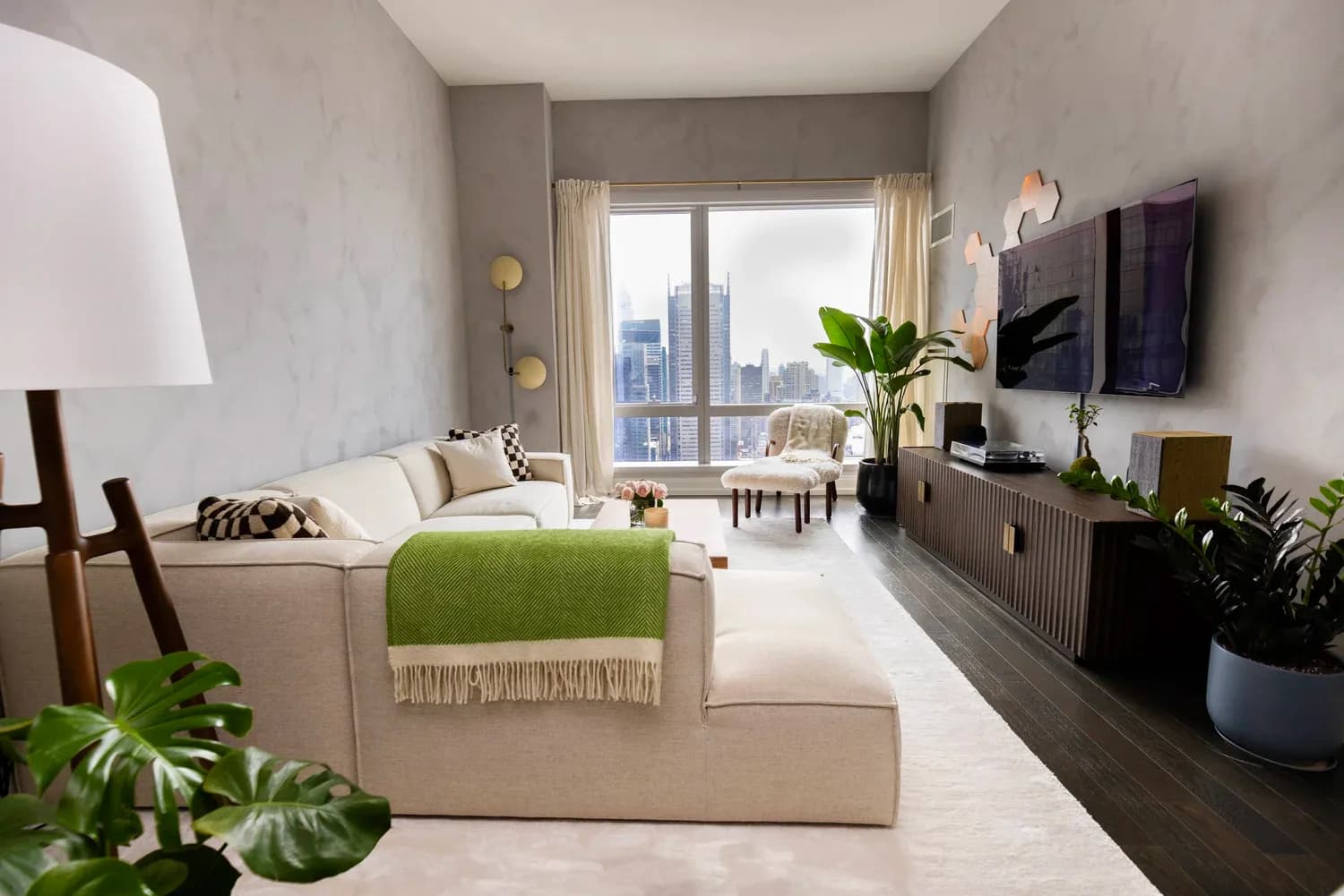 Modern living room with cream upholstered sofa, green throw blanket, dark media console, potted plants, and city skyline views through floor-to-ceiling windows by Matt Labombard.