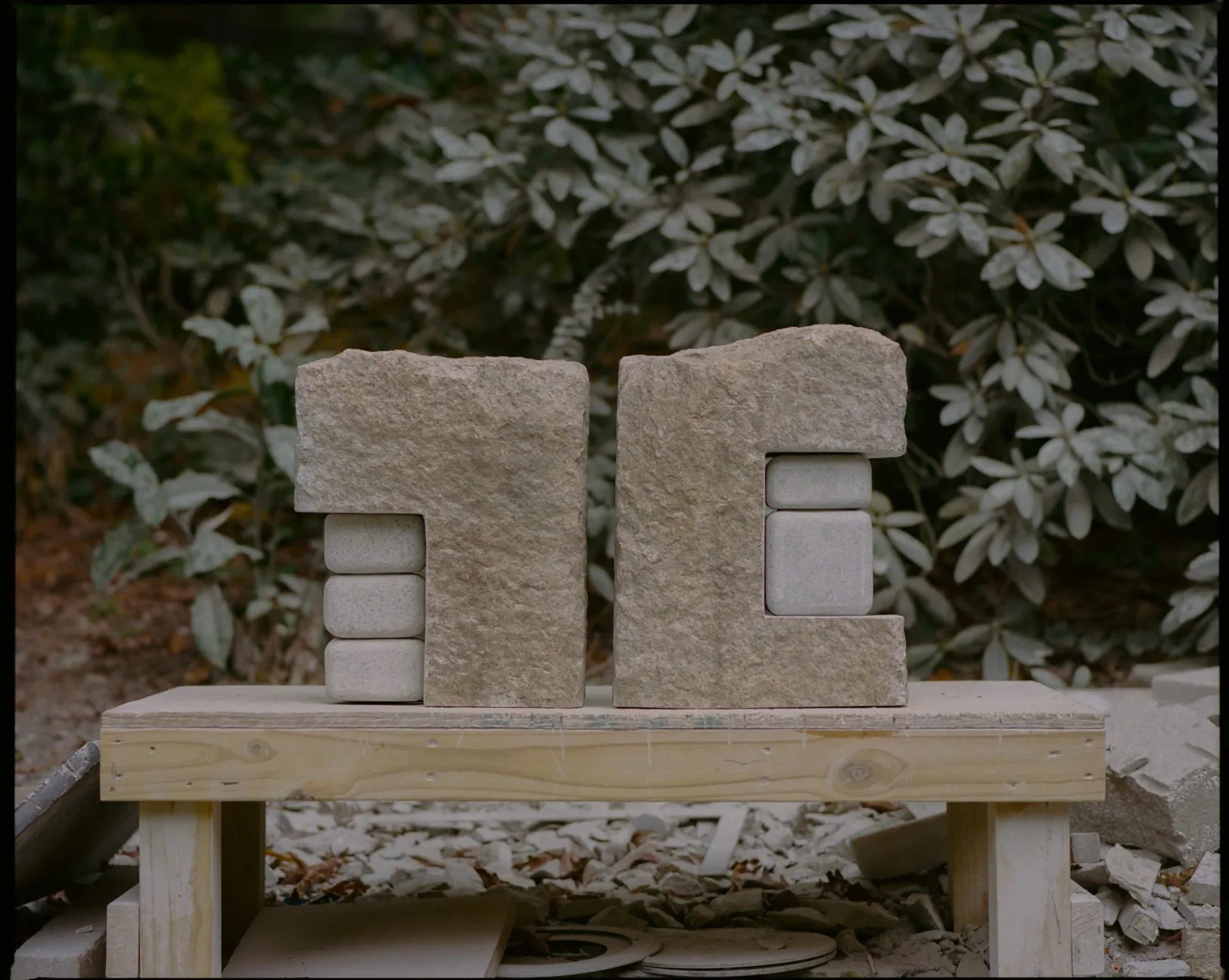 Stone sculptural installation featuring two rectangular forms with small square openings, mounted on a wooden base against foliage, by Matthew Byrd.