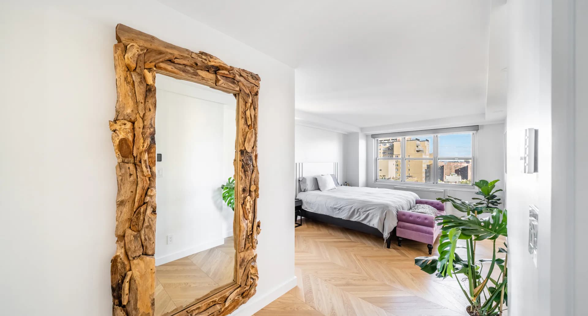 Modern bedroom with herringbone wood flooring, golden-framed mirror, and city views, designed by Matthew Ruopoli.