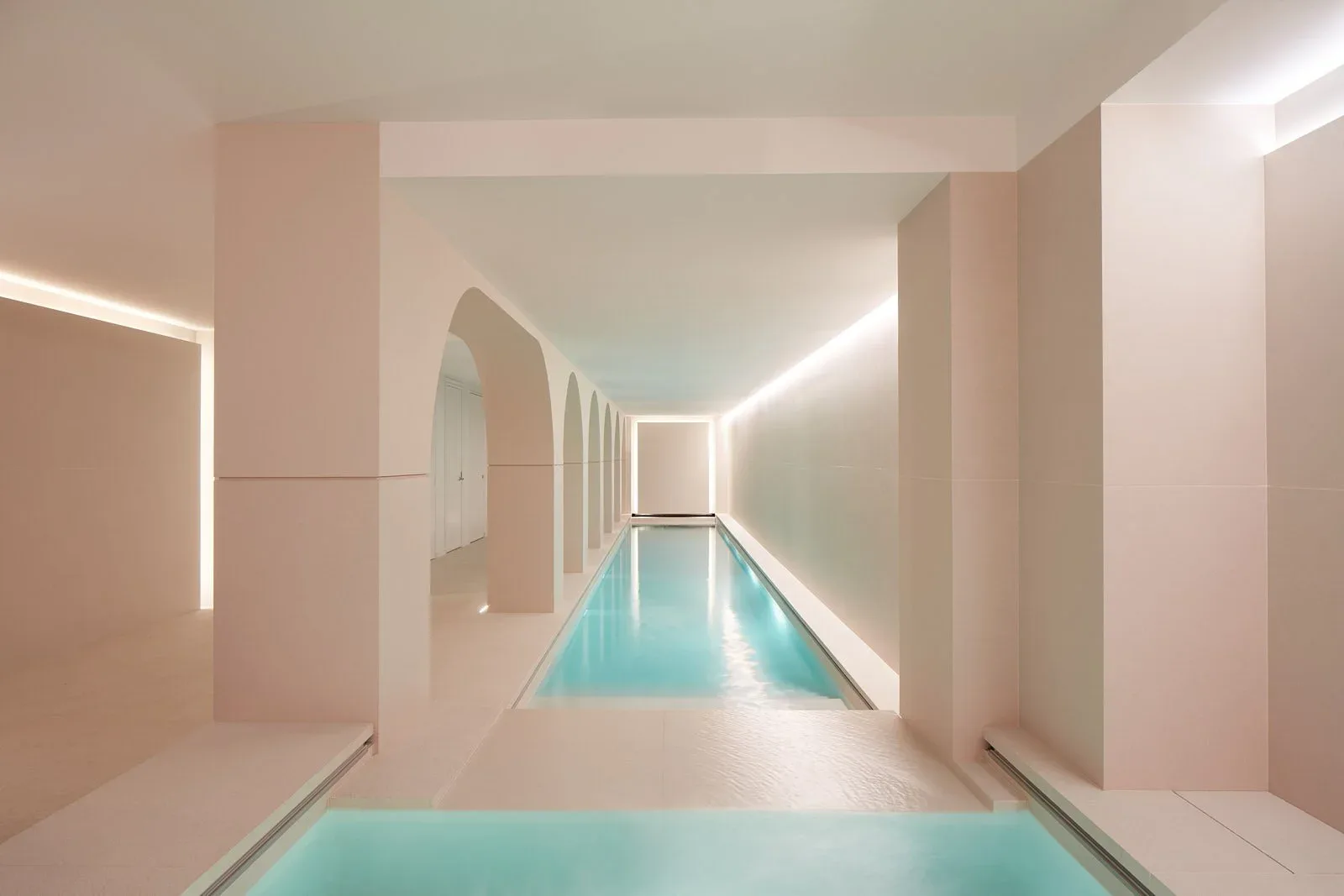 A minimalist indoor swimming pool with turquoise water flanked by beige walls and linear ceiling lights, designed by Matthew Tirschwell.