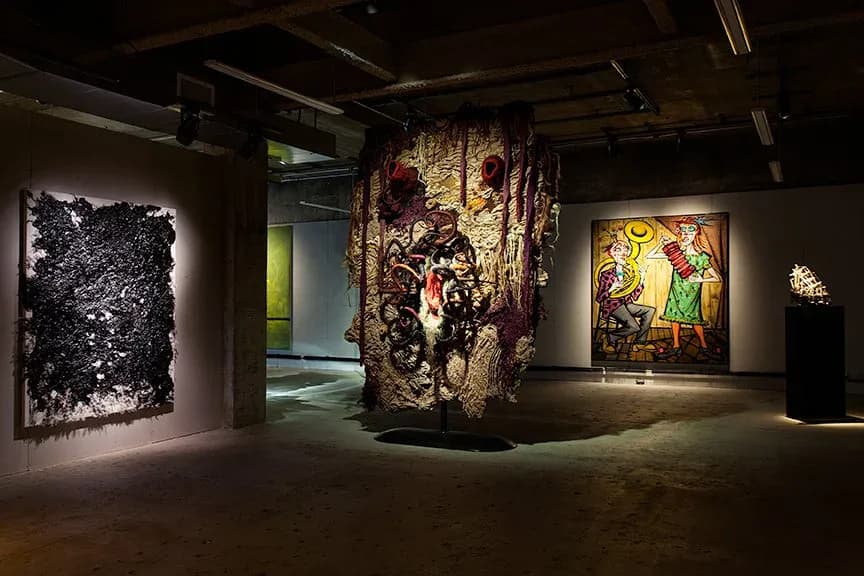 Contemporary art gallery with dark walls displaying vibrant abstract and figurative artworks, featuring a striking sculptural installation in the center, curated by Matthew Tirschwell.