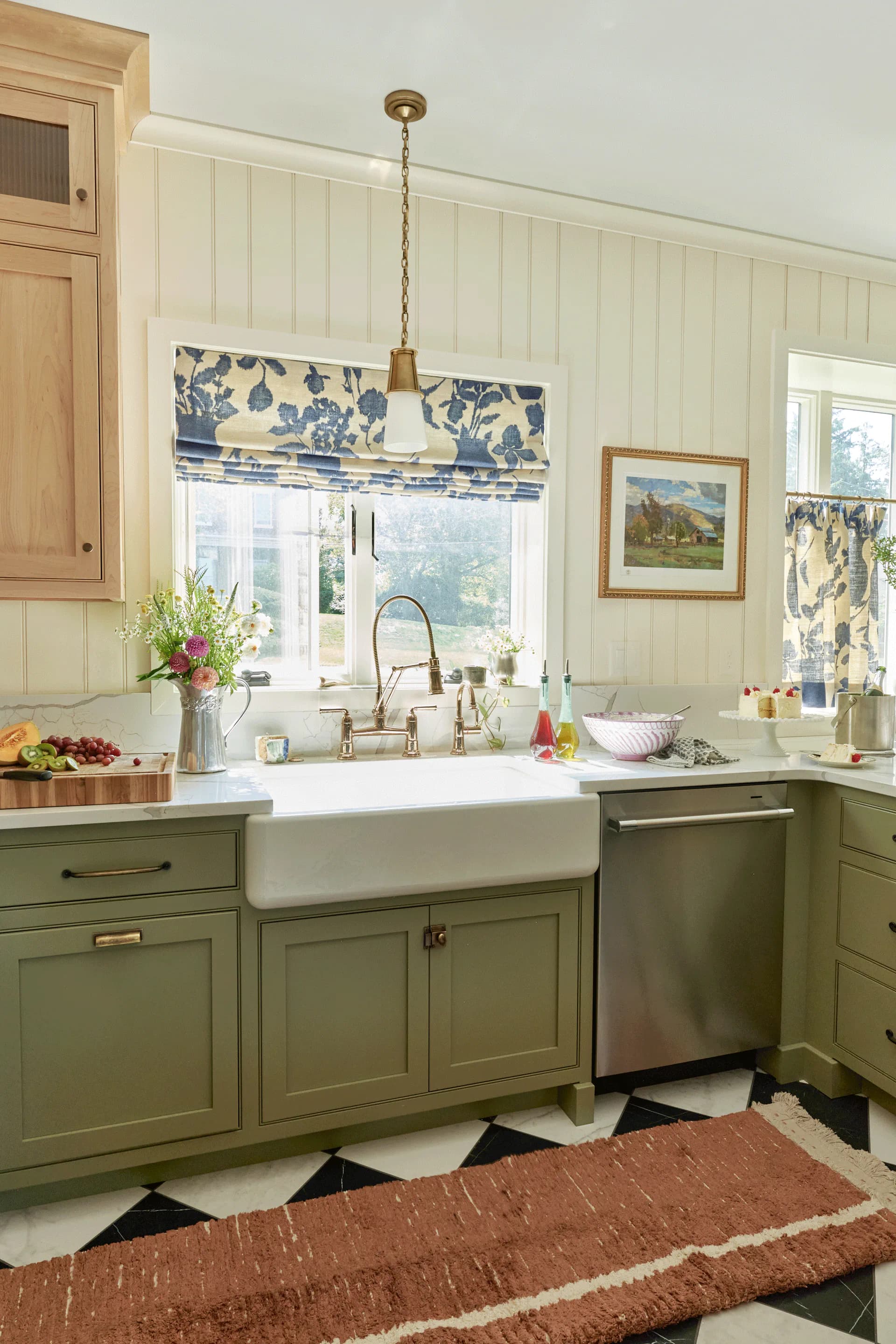A farmhouse kitchen with sage green cabinetry, white subway tile walls, white farmhouse sink, brass fixtures, and blue patterned window shade by Maura Clark.