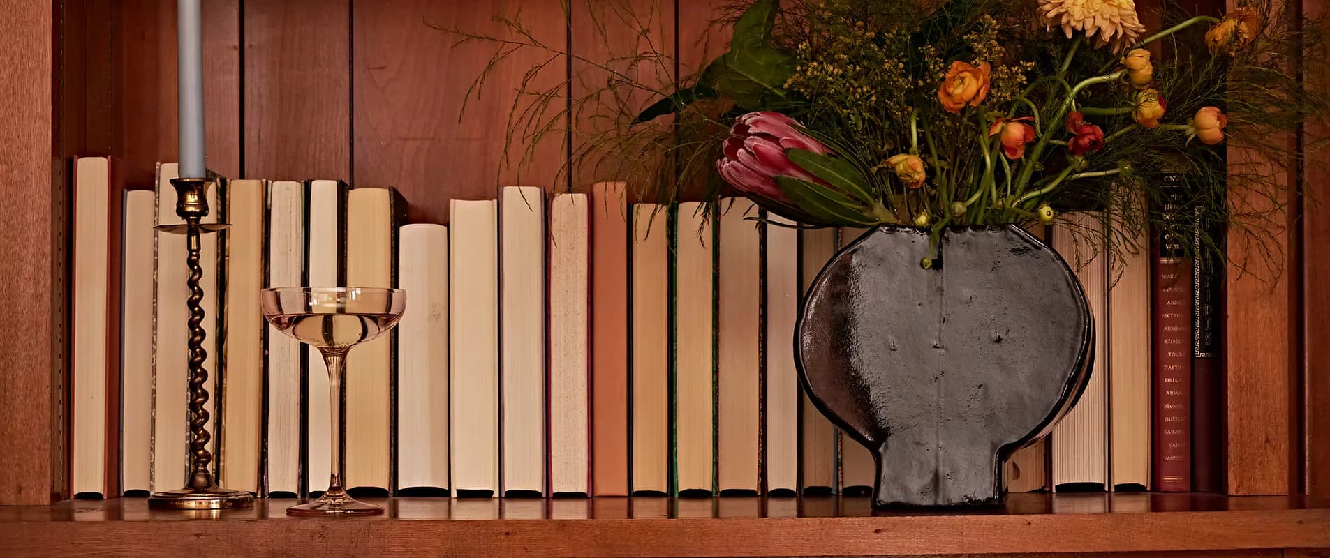 Warm-toned styled shelf display featuring cream candlesticks, decorative objects, and a charcoal vase with fresh flowers against a terracotta wall by Maura Clark.