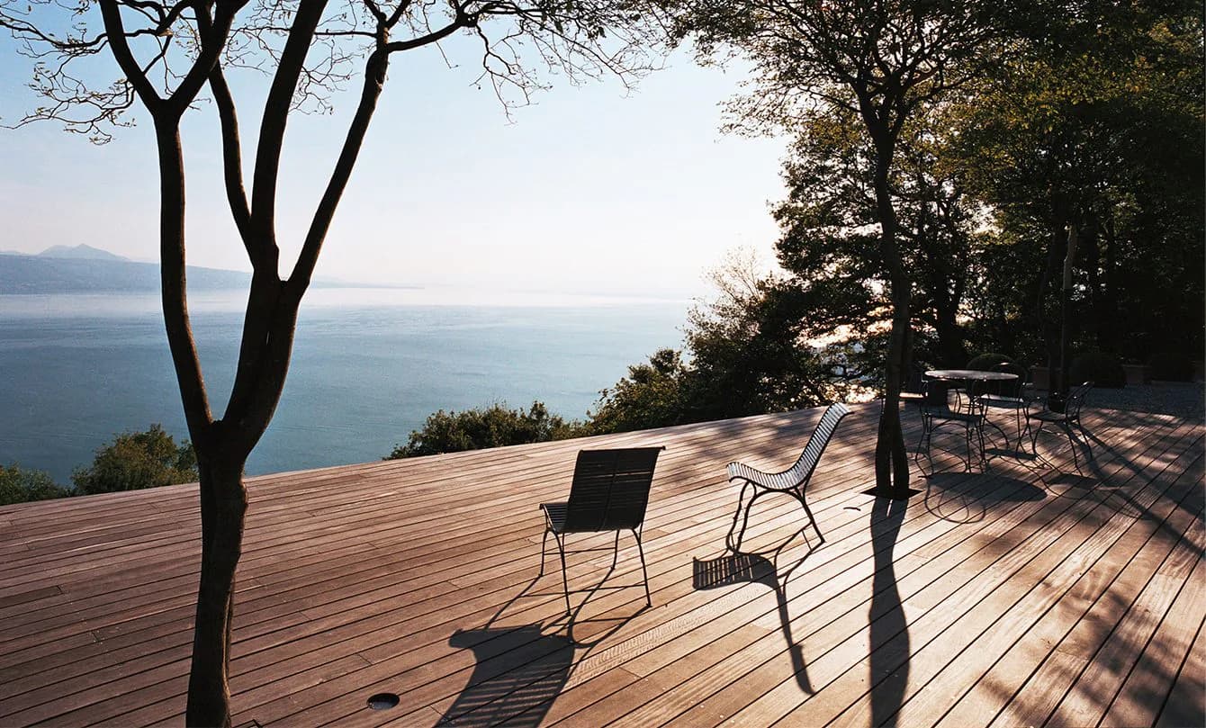 Waterfront terrace with wooden decking, modern seating, and mature trees framing a serene lake view, designed by Maya Alawa.