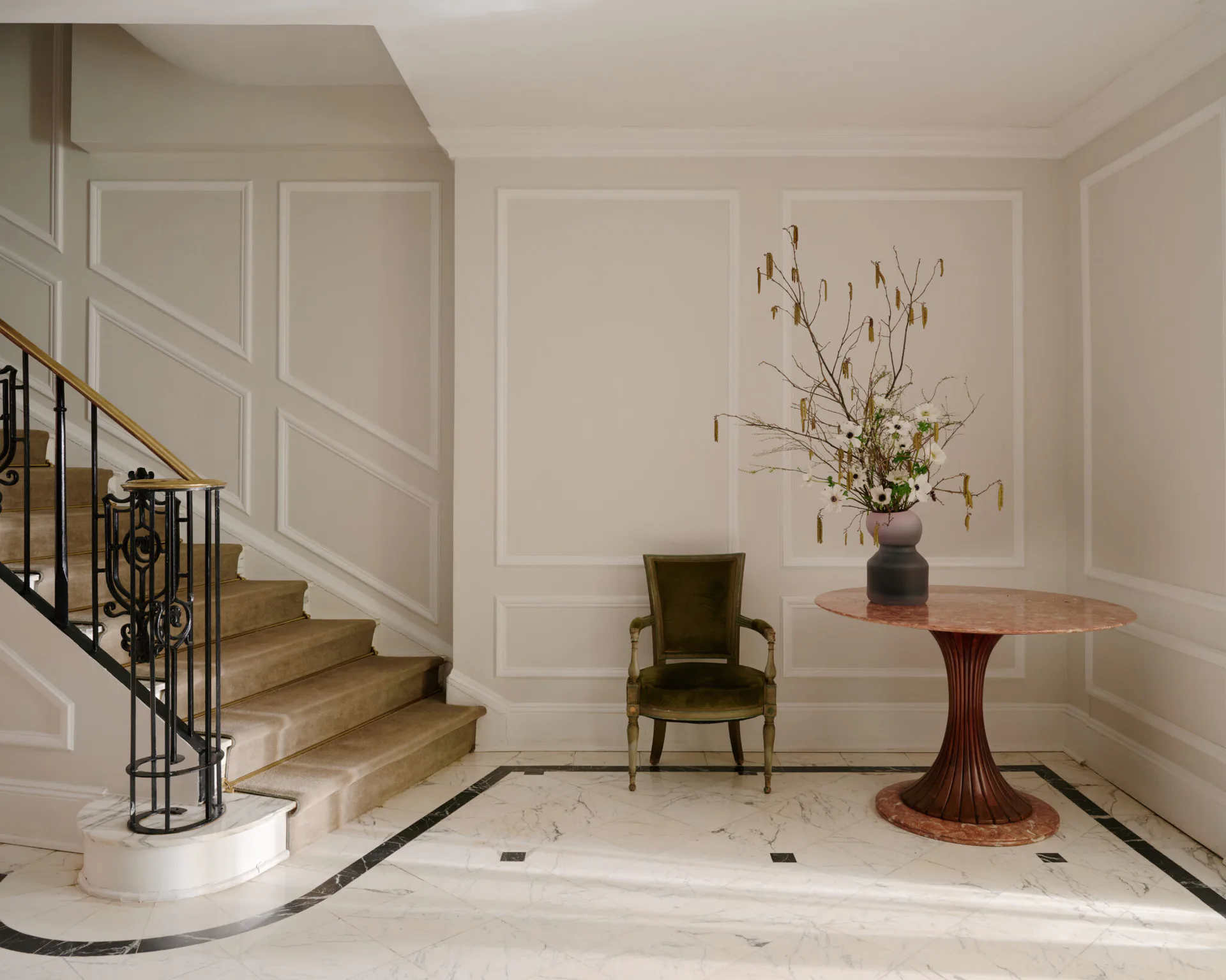 Minimalist foyer with curved staircase, olive chair, round wood table with dried florals, white paneled walls, and polished marble floor by Maylene Reyes.