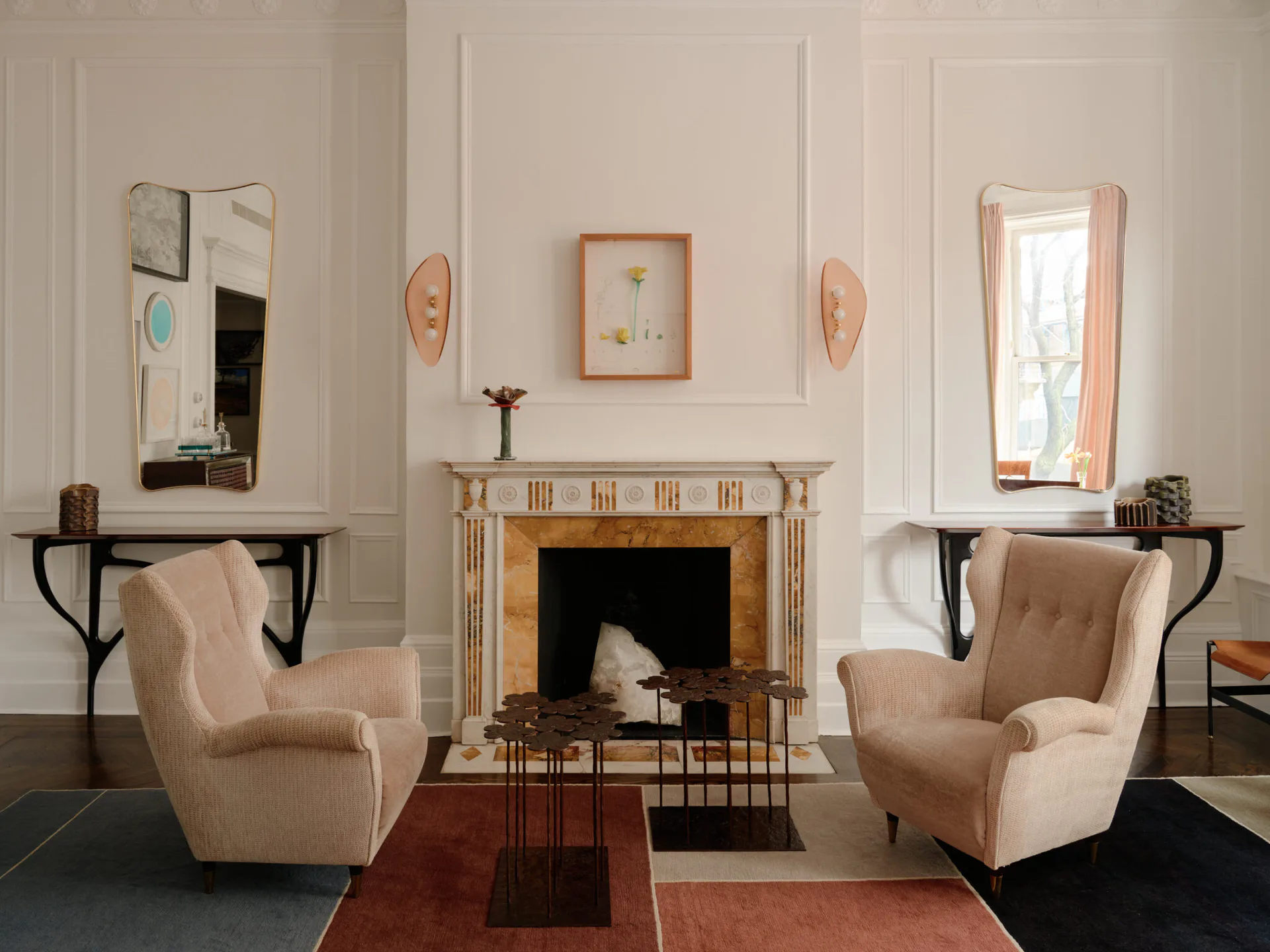 Elegant living room with symmetrical beige armchairs flanking a fireplace, mirrors and sconces on cream walls, terracotta area rug, designed by Maylene Reyes.