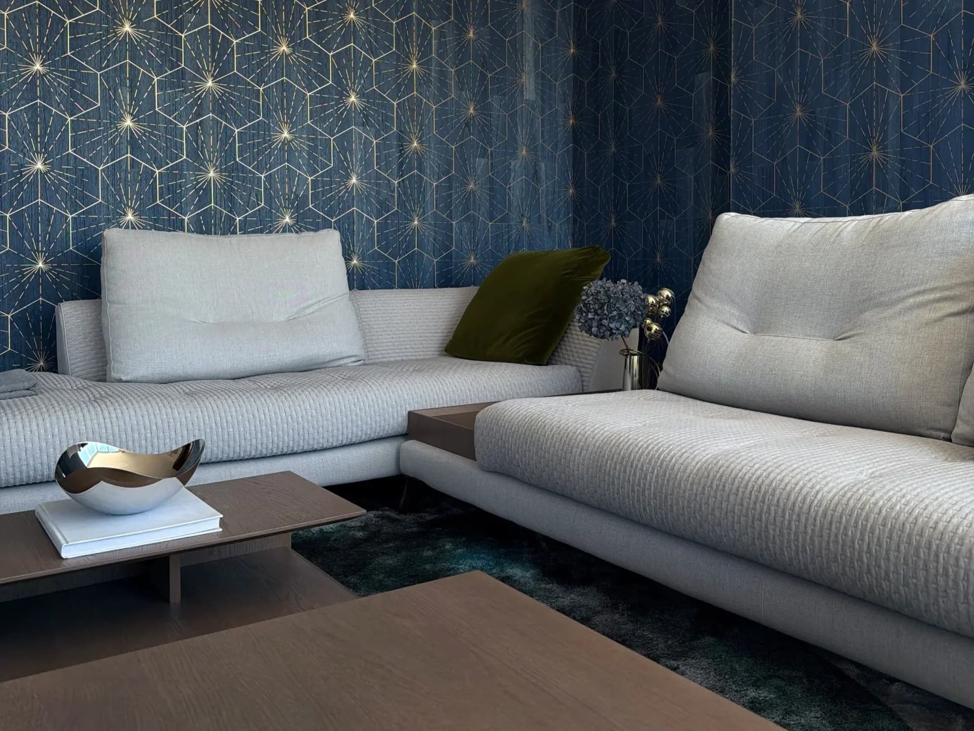Modern living room with light gray sectional sofa, dark teal geometric patterned wallpaper, wood coffee table, and decorative accents by Mazen Eshak.