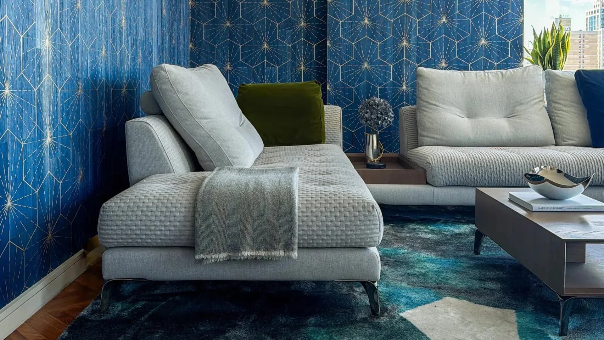Modern living room interior with geometric patterned blue wallpaper, light gray upholstered seating, teal area rug, and minimalist furnishings by Mazen Eshak.