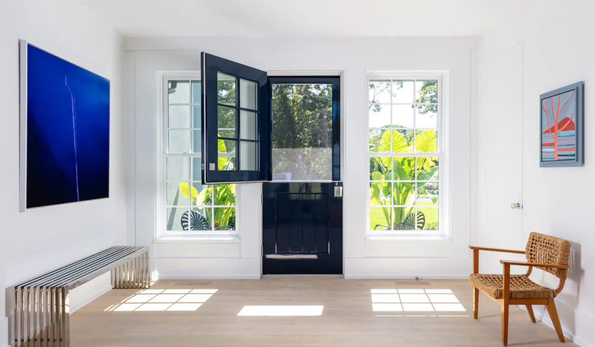 Minimalist interior with white walls, light wood flooring, and floor-to-ceiling windows opening to greenery, featuring a black accent door, blue artwork, and mid-century modern furnishings by Meagan Maudsley.