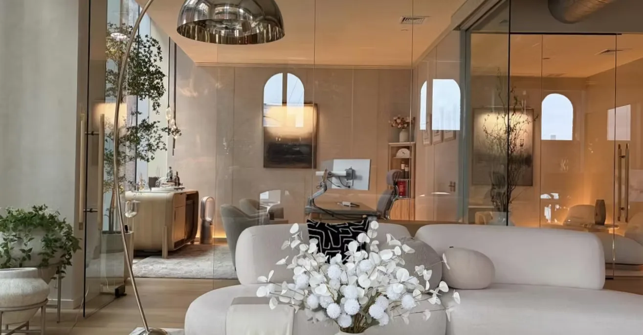 Luxurious modern living room with white curved sofa, white floral arrangement, glass partition, warm ambient lighting, and neutral palette by Megen Hlavacek.