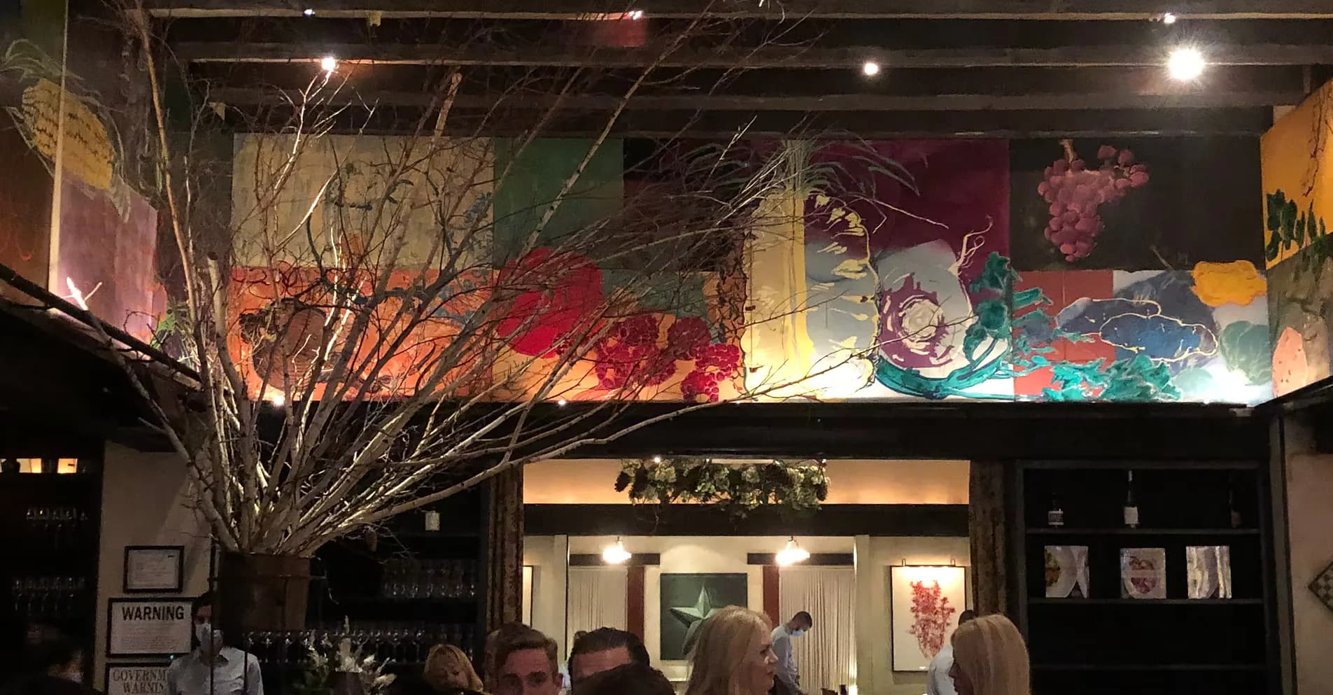 Vibrant storefront interior with colorful abstract mural installation, bare branch sculptural elements, and warm ambient lighting by Megen Hlavacek.