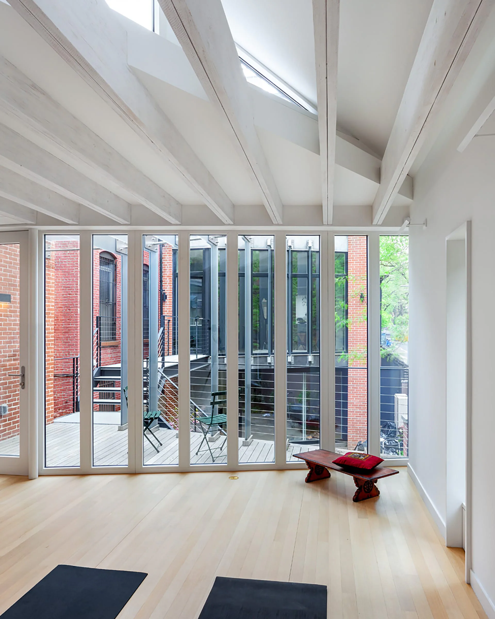 Bright modern interior with white exposed beam ceiling, light wood flooring, and expansive glass doors opening to a brick terrace, designed by Meghan Laky.
