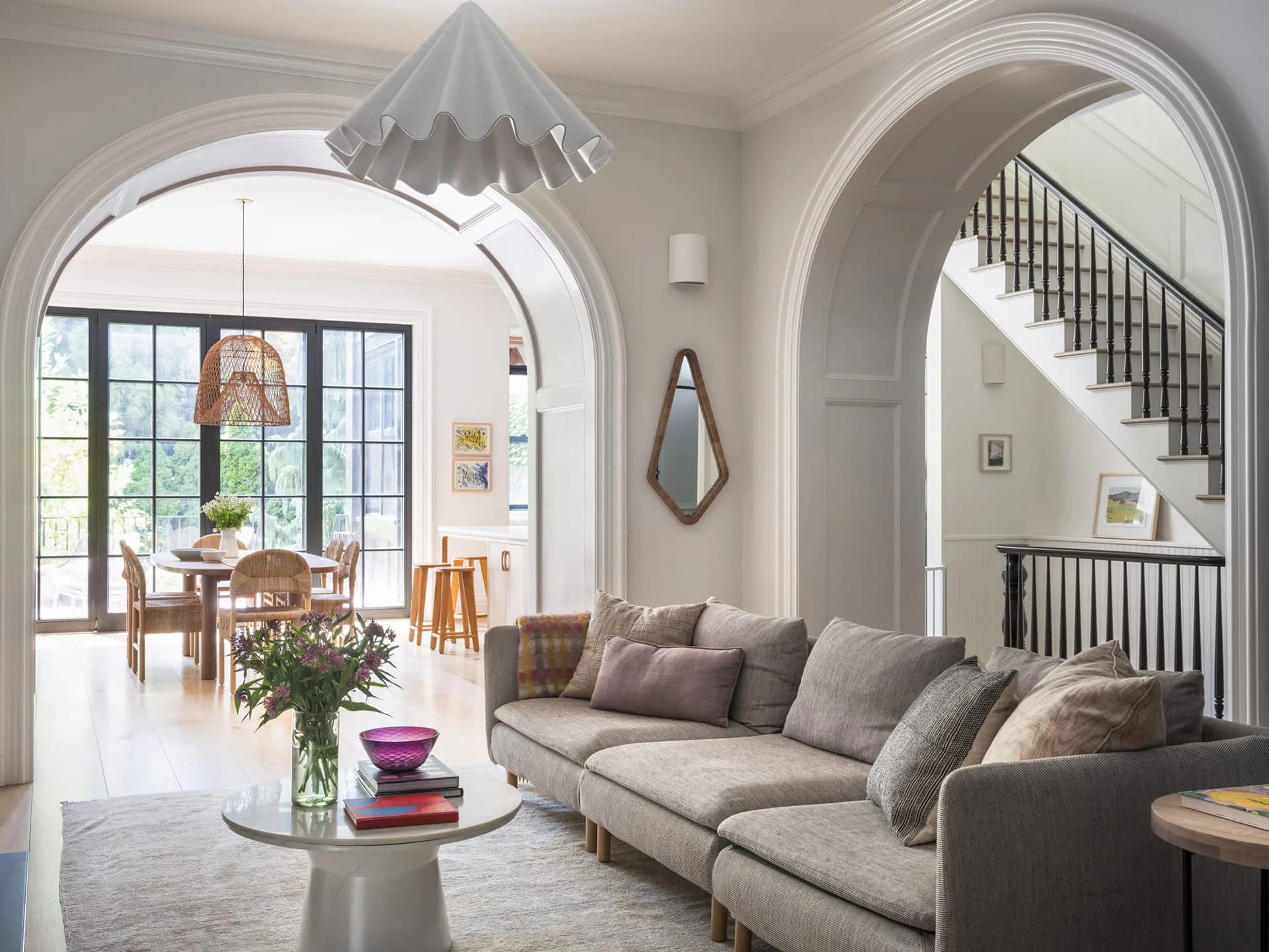 Bright modern living room with gray sectional sofa, arched doorways, geometric pendant light, and views to dining area by Meghan Laky.