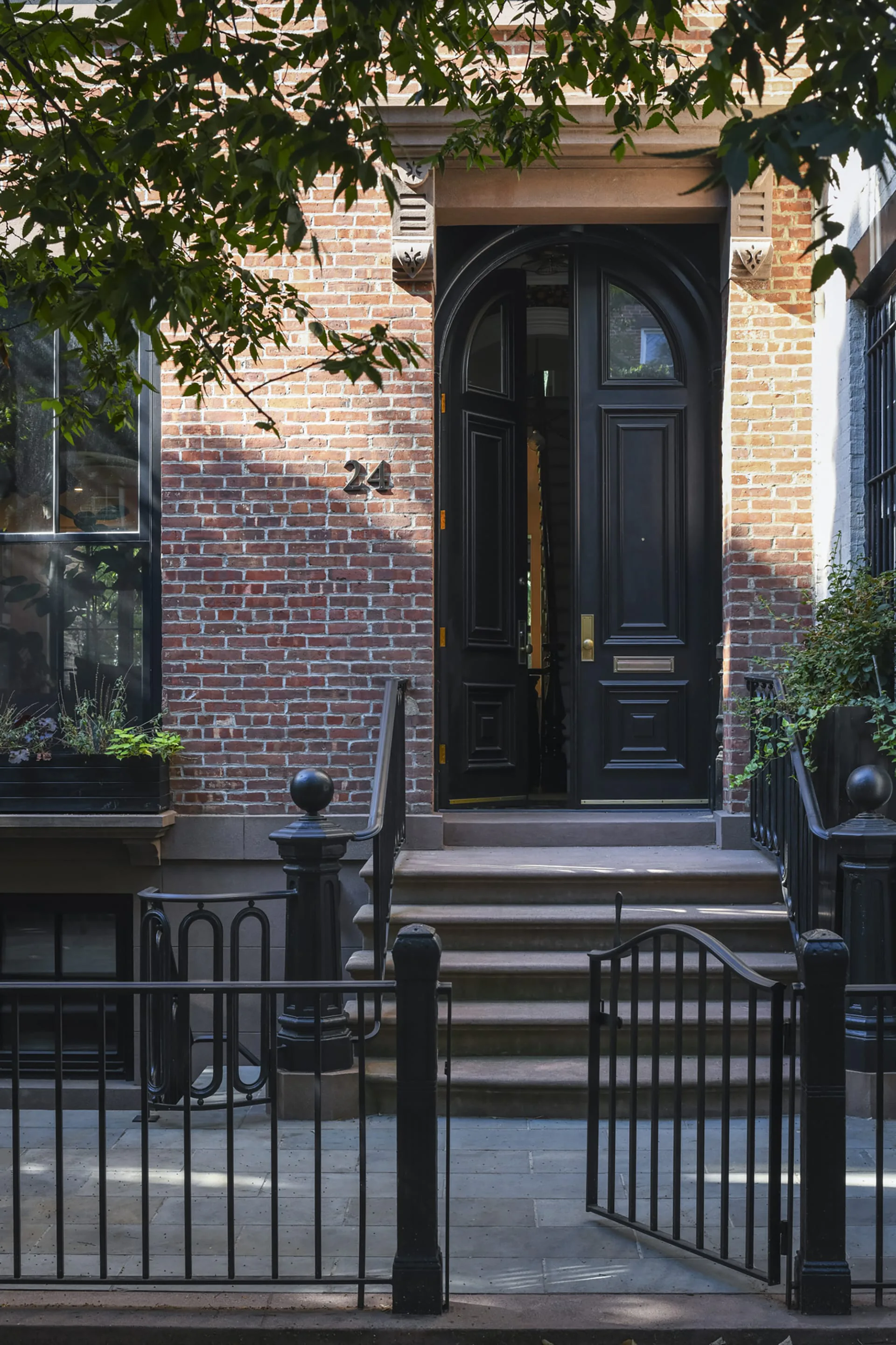 A black arched entrance door on a brick facade with iron railings and steps, featuring brass hardware and surrounded by climbing ivy by Meghan Laky.