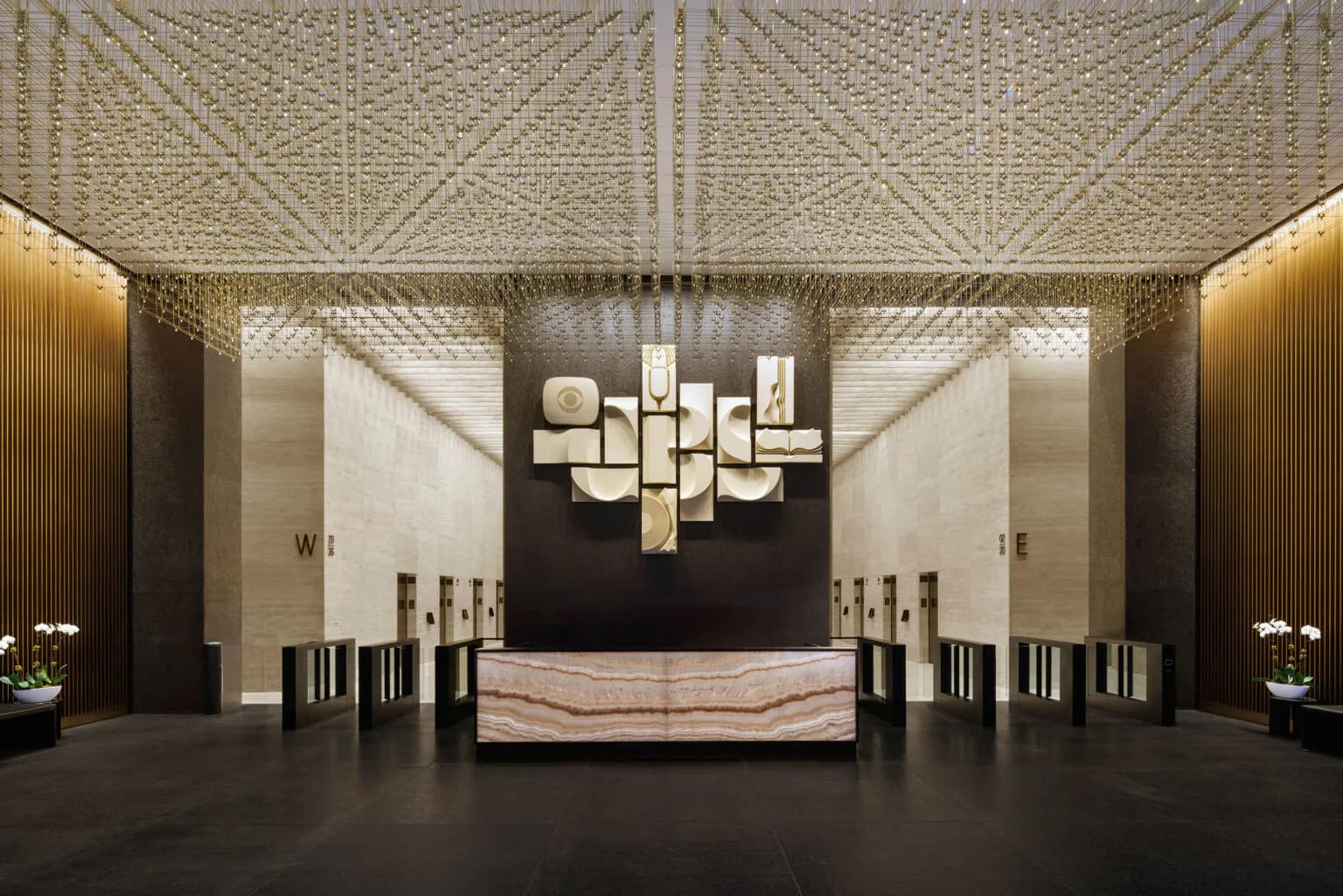 Modern luxury lobby with geometric patterned ceiling, dark accent wall, geometric sculptural chandelier, marble reception desk, and symmetrical wood-paneled side walls by Mel Casipit.