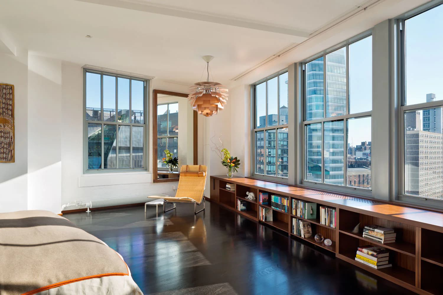 Bright modern bedroom with floor-to-ceiling windows overlooking a city skyline, dark wood built-in shelving, pendant lighting, and minimalist furnishings by Melanie Freundlich.