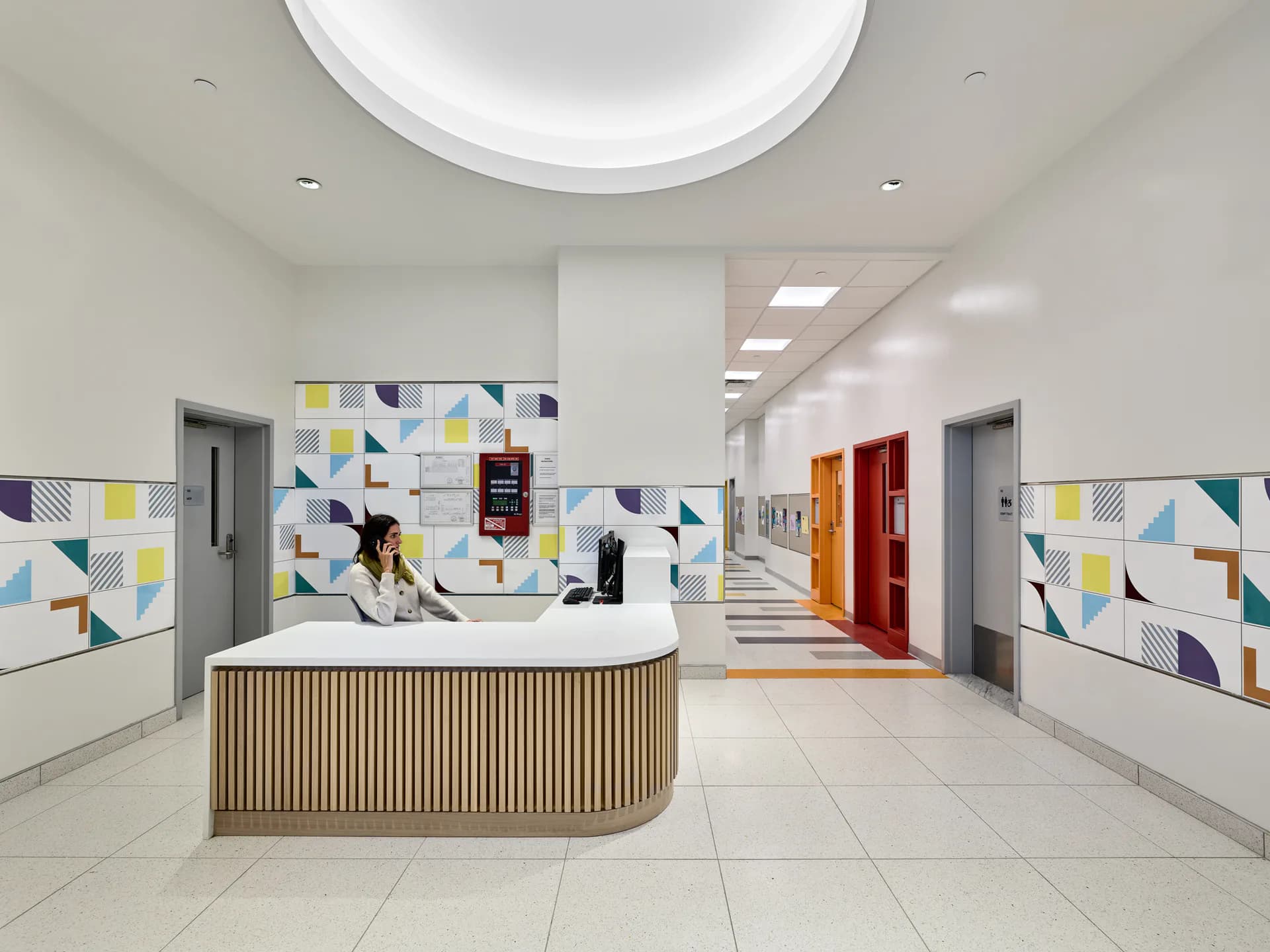 Modern medical clinic reception area with curved wooden-paneled desk, geometric tile wall mural in blue, yellow, and pink, skylights, and corridor access to red doors, designed by Melanie Freundlich.