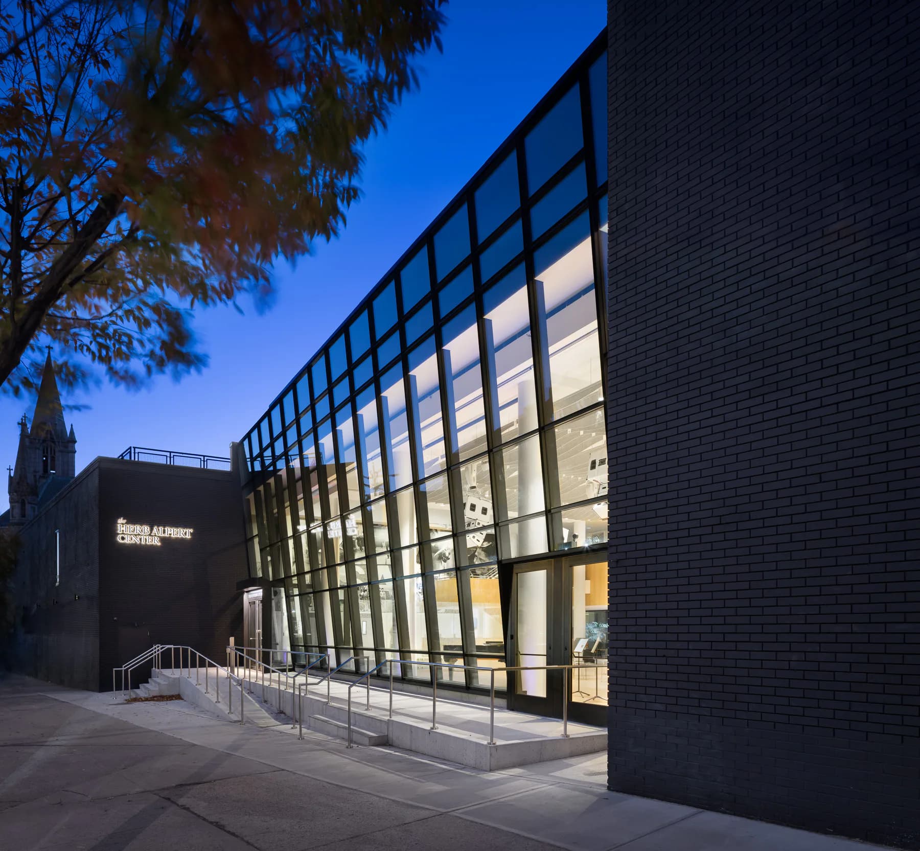 Modern brick and glass cultural center photographed at dusk, featuring illuminated gridded windows and dark masonry exterior by Melanie Freundlich.