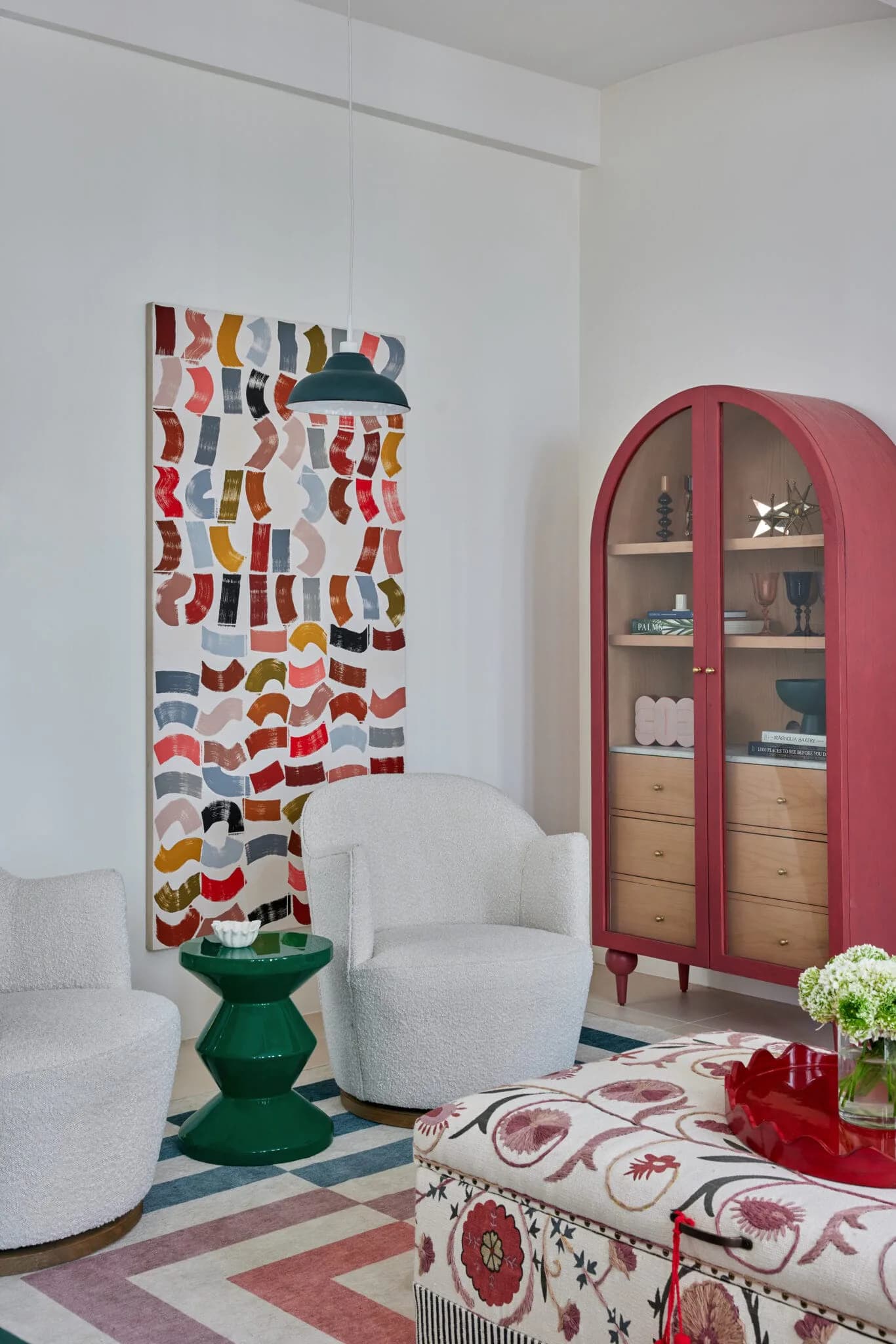 A modern living room interior featuring white curved chairs, a red arched cabinet, colorful patterned textiles, and green pedestal accent table designed by Melanie King.