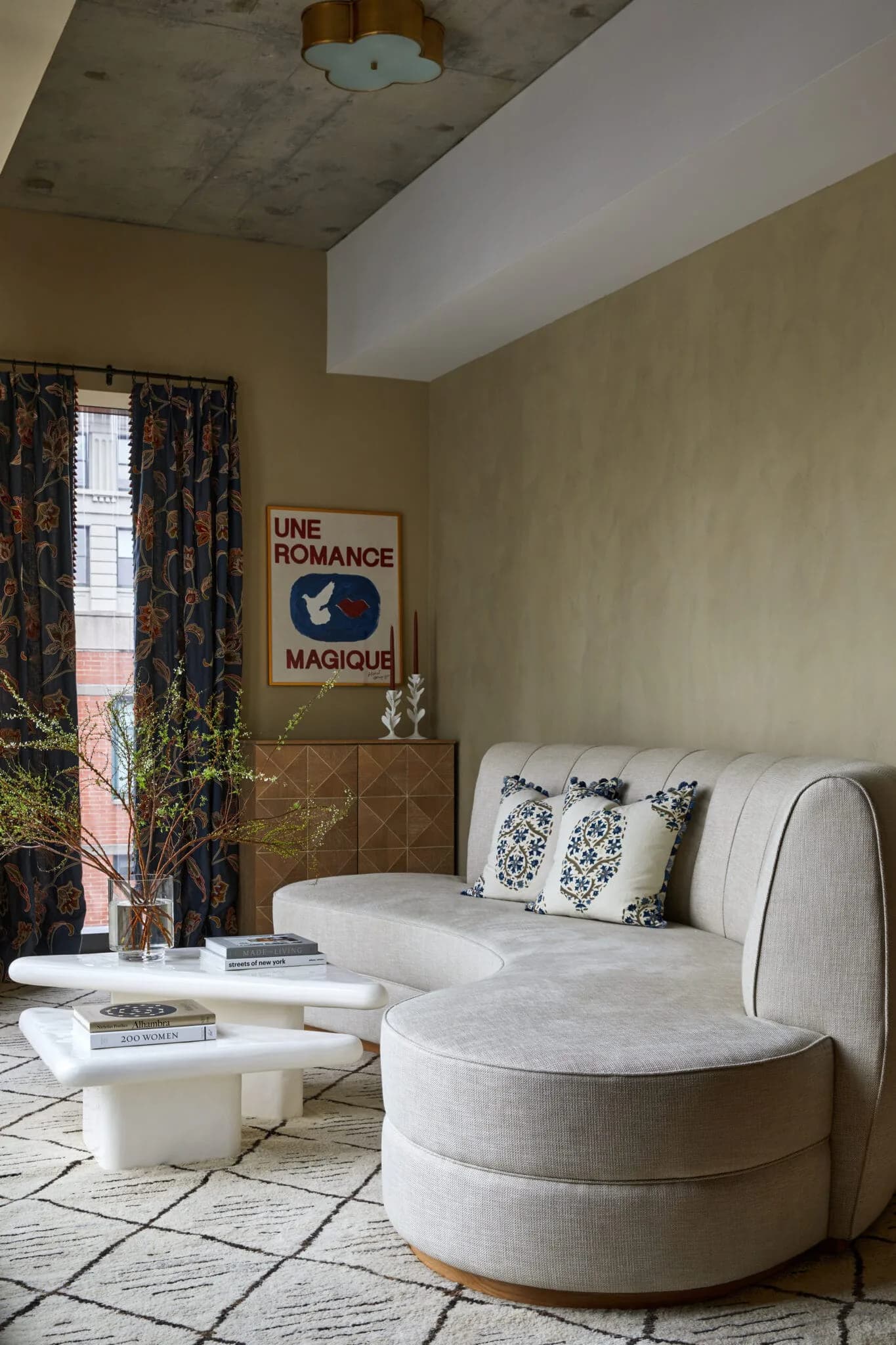 Modern living room with curved cream sectional sofa, white sculptural coffee table, sage green walls, patterned curtains, and vintage poster by Melanie King.