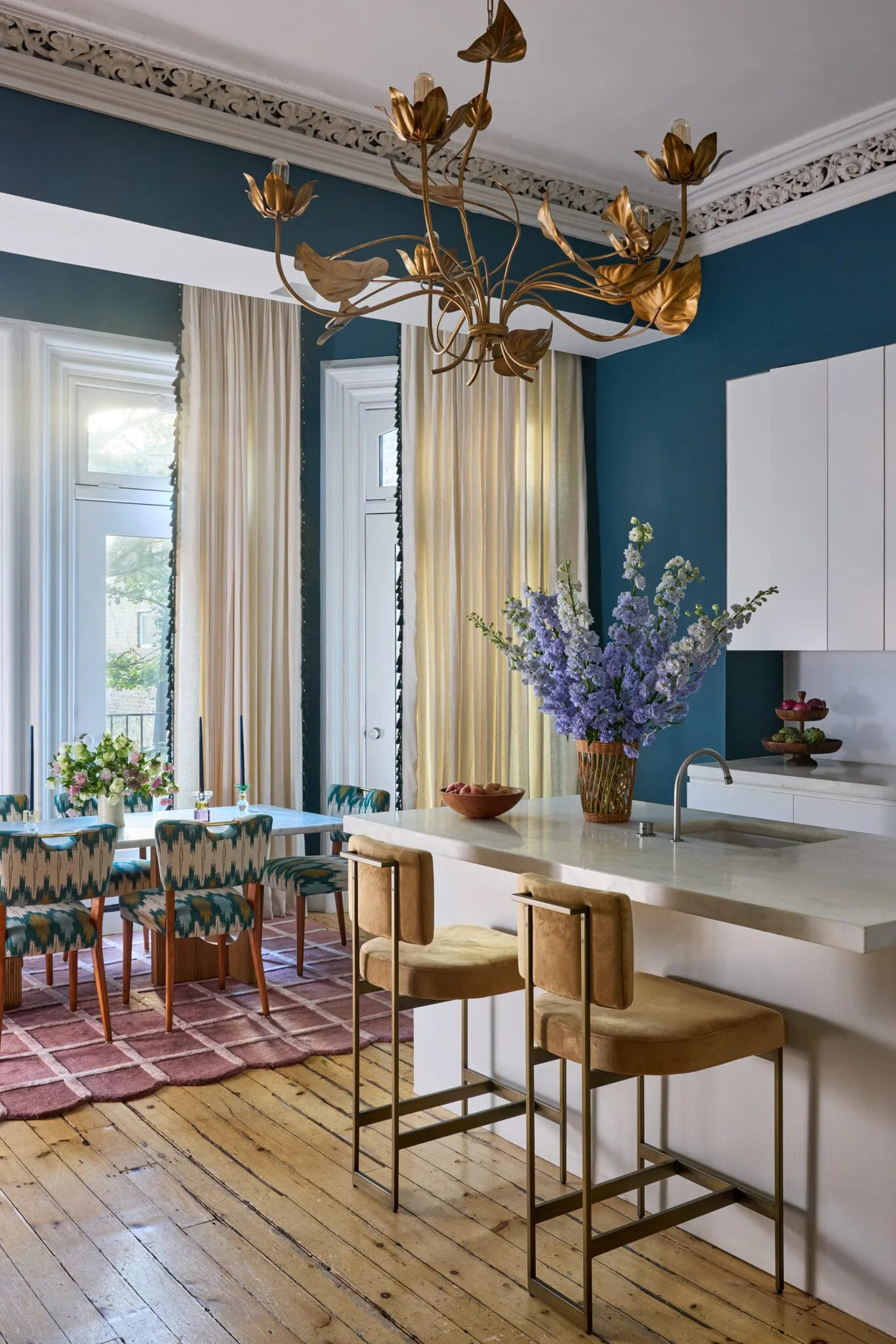 Contemporary dining room with deep teal walls, brass floral chandelier, white cabinetry, and cream curtains, designed by Melanie King.