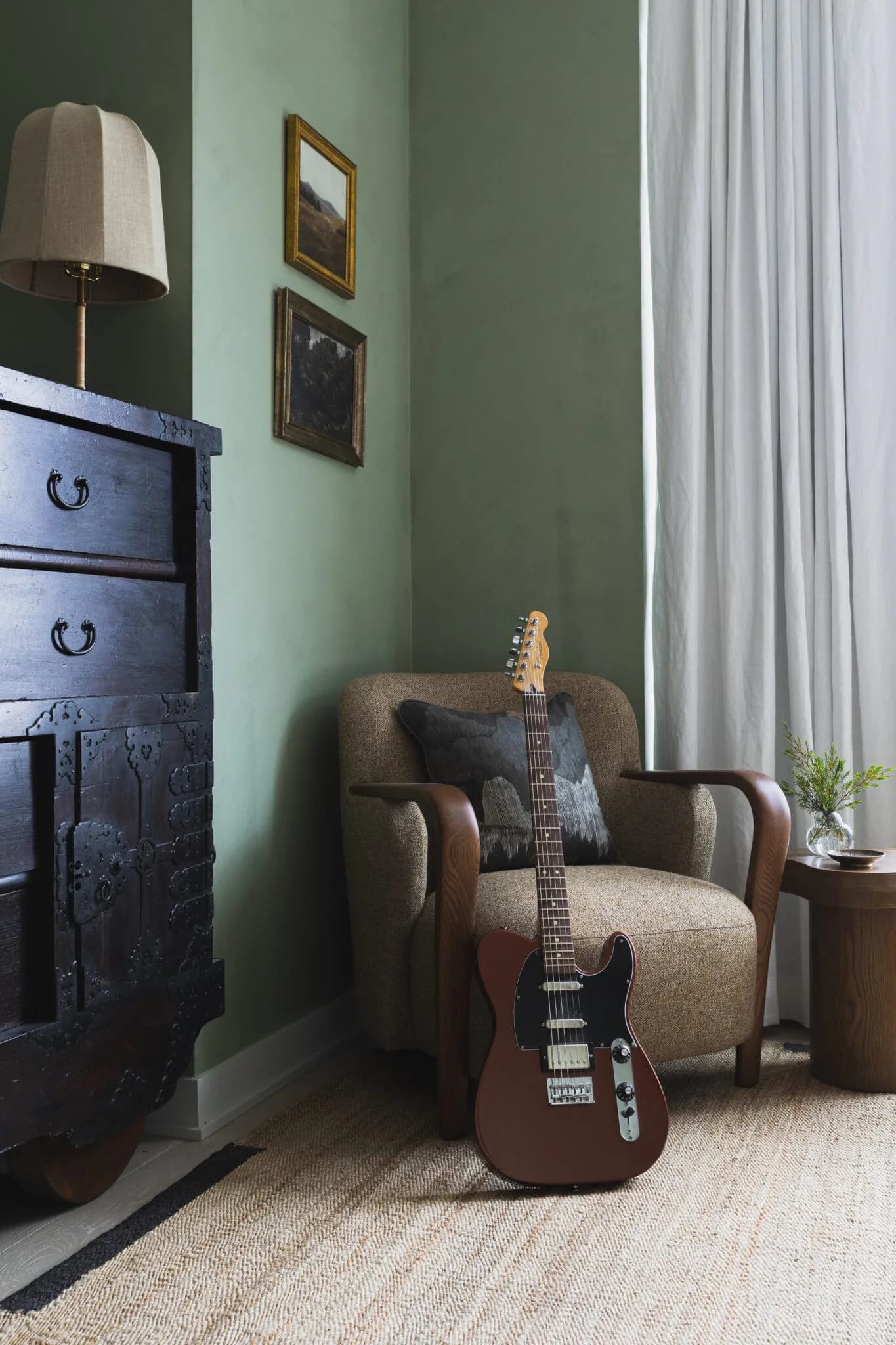 A sage green bedroom corner featuring a taupe upholstered armchair with a guitar leaning against it, a dark navy dresser with brass hardware, and framed artwork, designed by Melanie King.