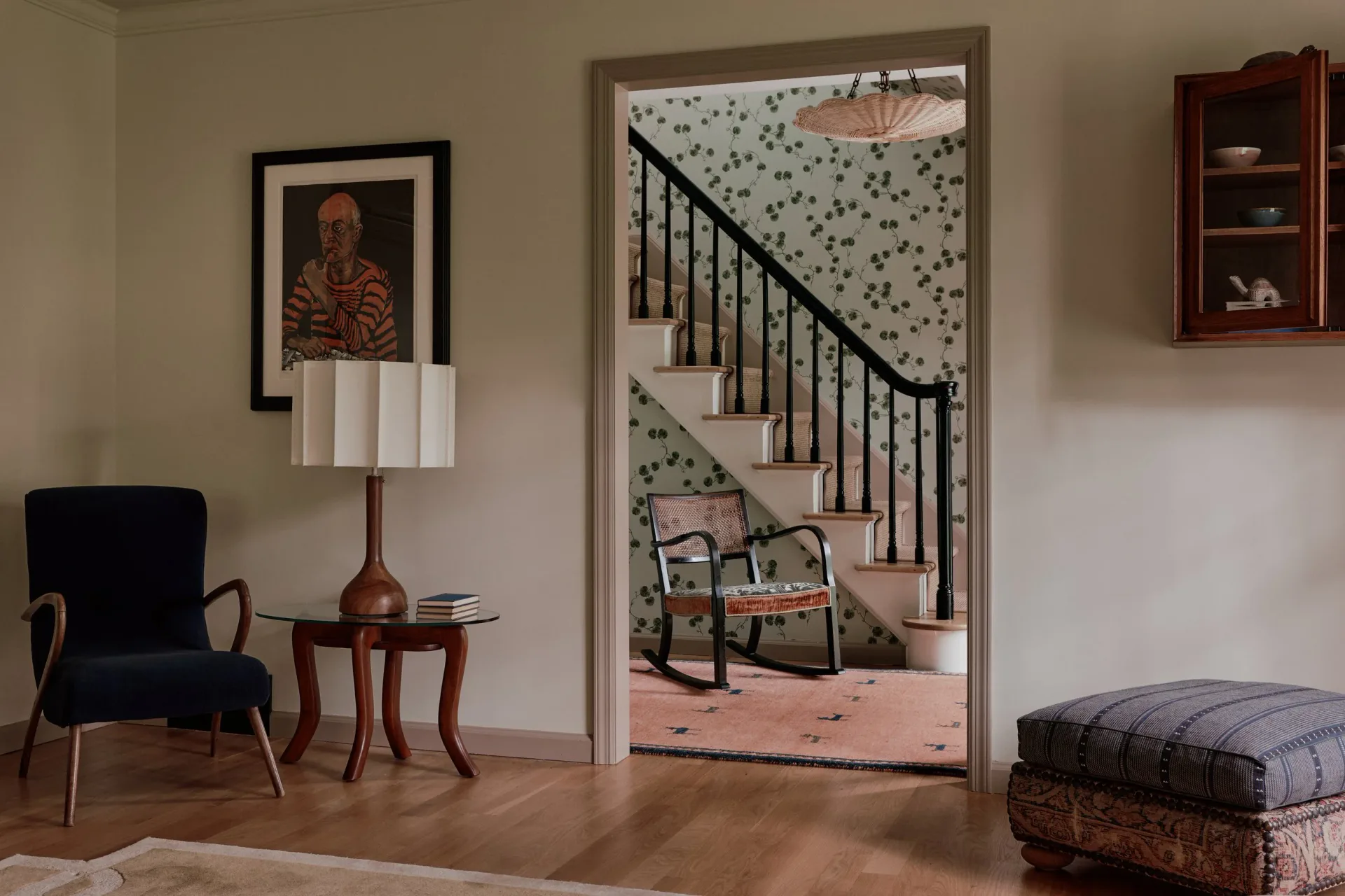 Modern interior space with black chair, wooden side table, and staircase visible through doorway with patterned wallpaper, designed by Melissa Lee.