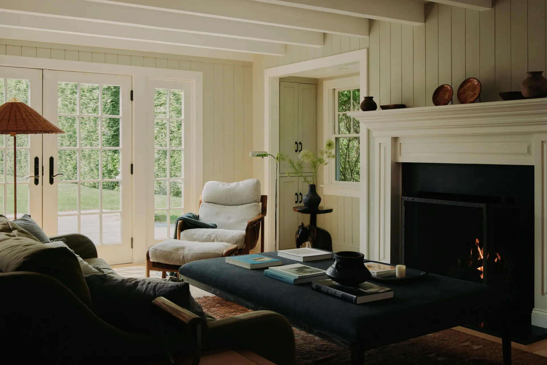 Elegant living room with cream and green palette, featuring a fireplace, large windows, dark furniture, and classic architectural details by Melissa Lee.