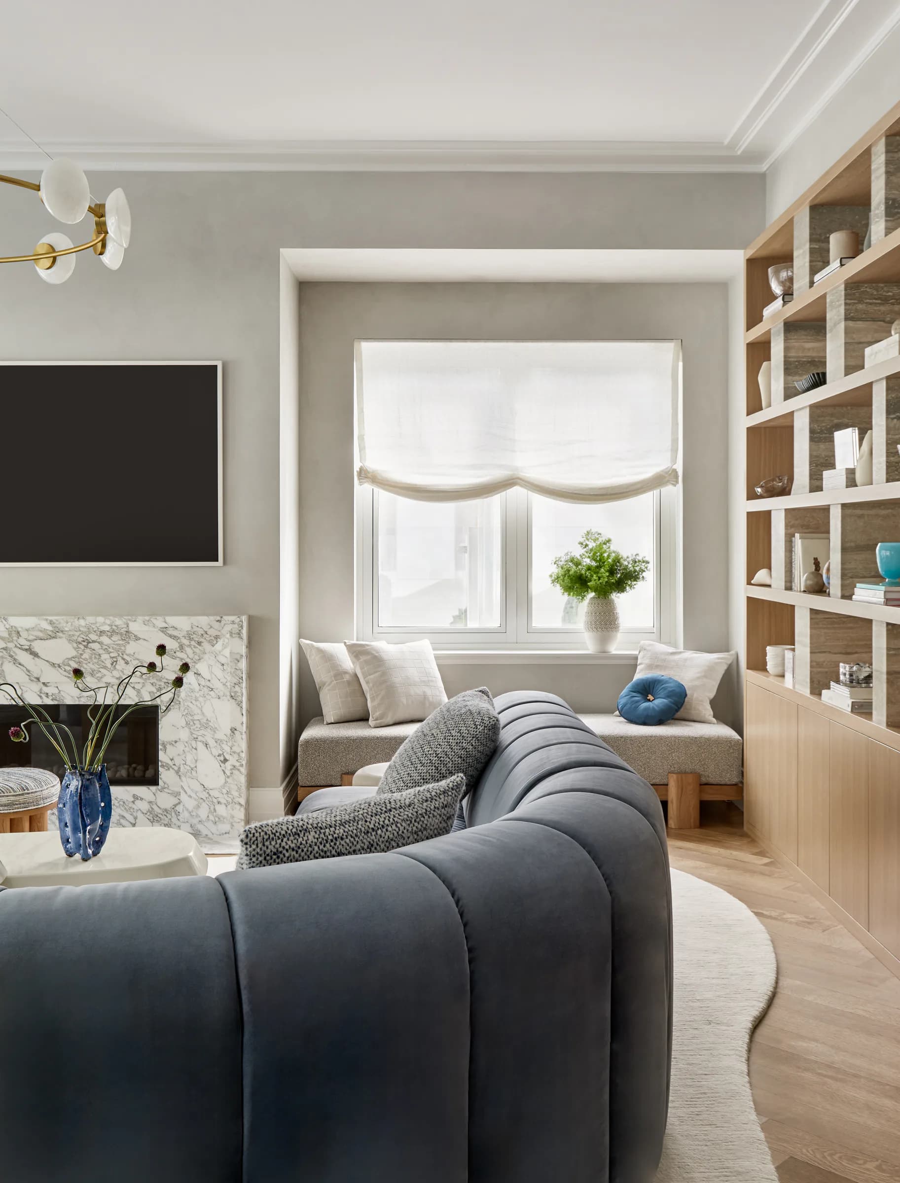 Contemporary living room with gray walls, marble fireplace, dark curved sofa, wooden shelving, and bright window seat by Melissa McAlister.