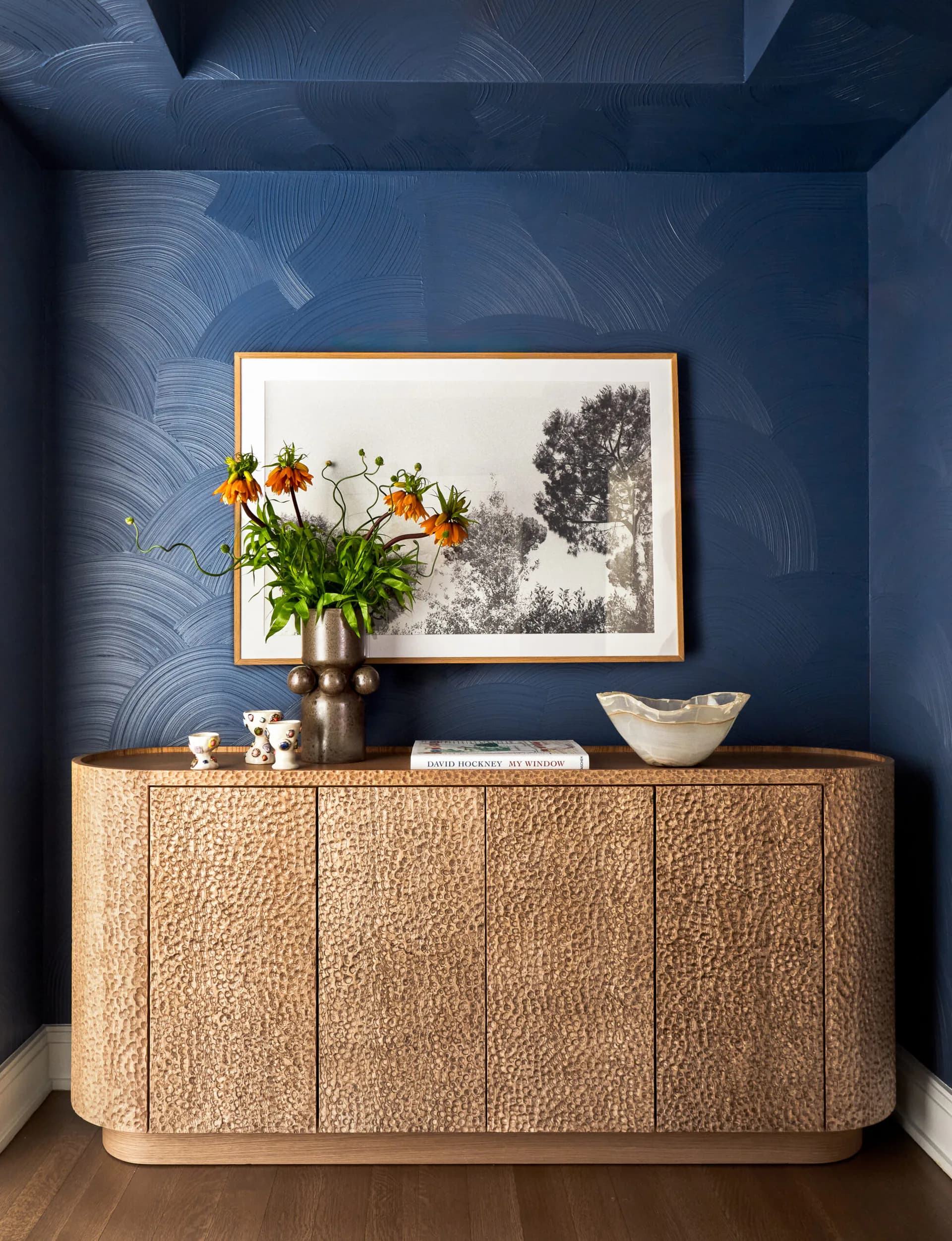 Dark blue accent wall with textured finish frames a natural fiber credenza topped with a brass vase of yellow flowers and framed landscape art by Melissa McAlister.