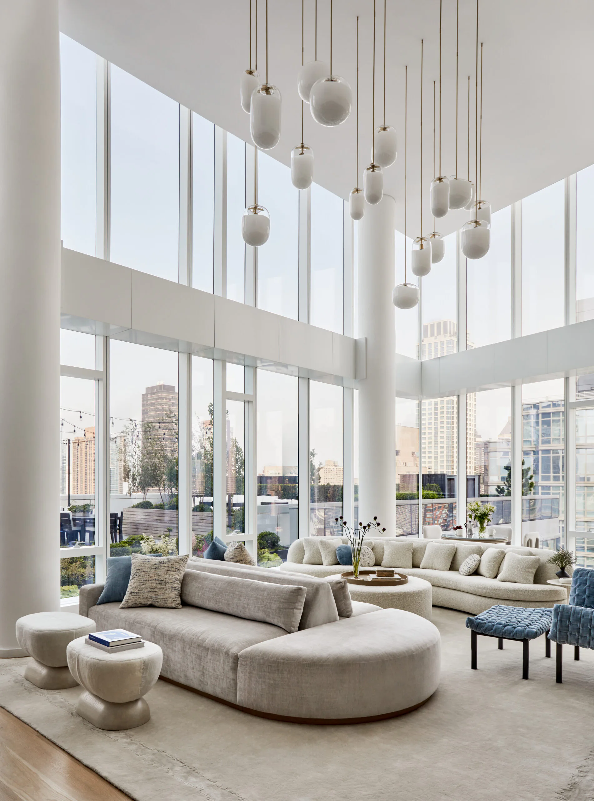 A luxurious double-height living room with curved modular seating, cascading pendant lights, floor-to-ceiling windows overlooking a city skyline, and a neutral palette of cream and beige, by Melissa McAlister.