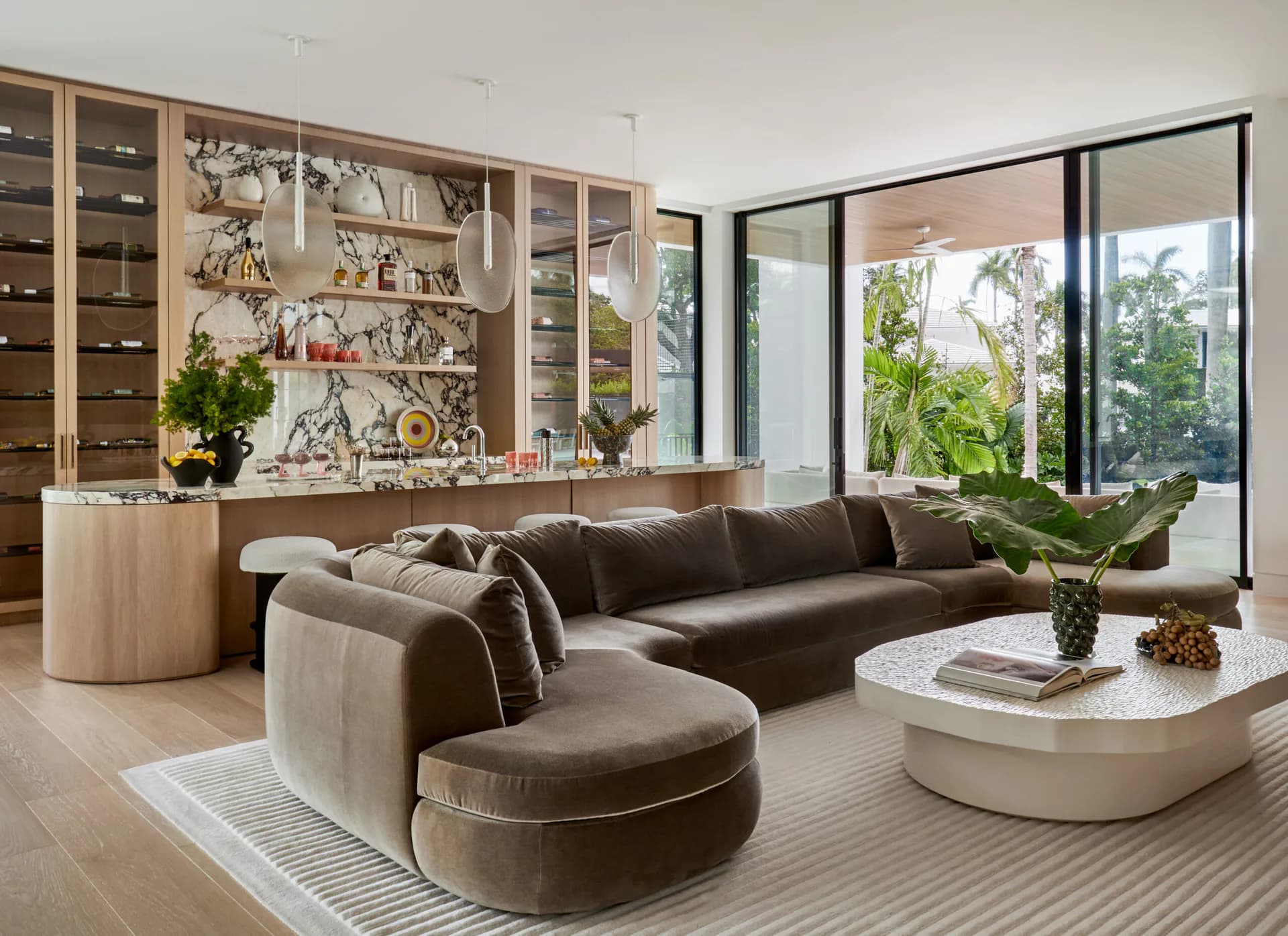 Modern living room with curved sectional sofa in taupe velvet, curved ottoman, white marble coffee table, and floor-to-ceiling windows overlooking greenery by Melissa McAlister.