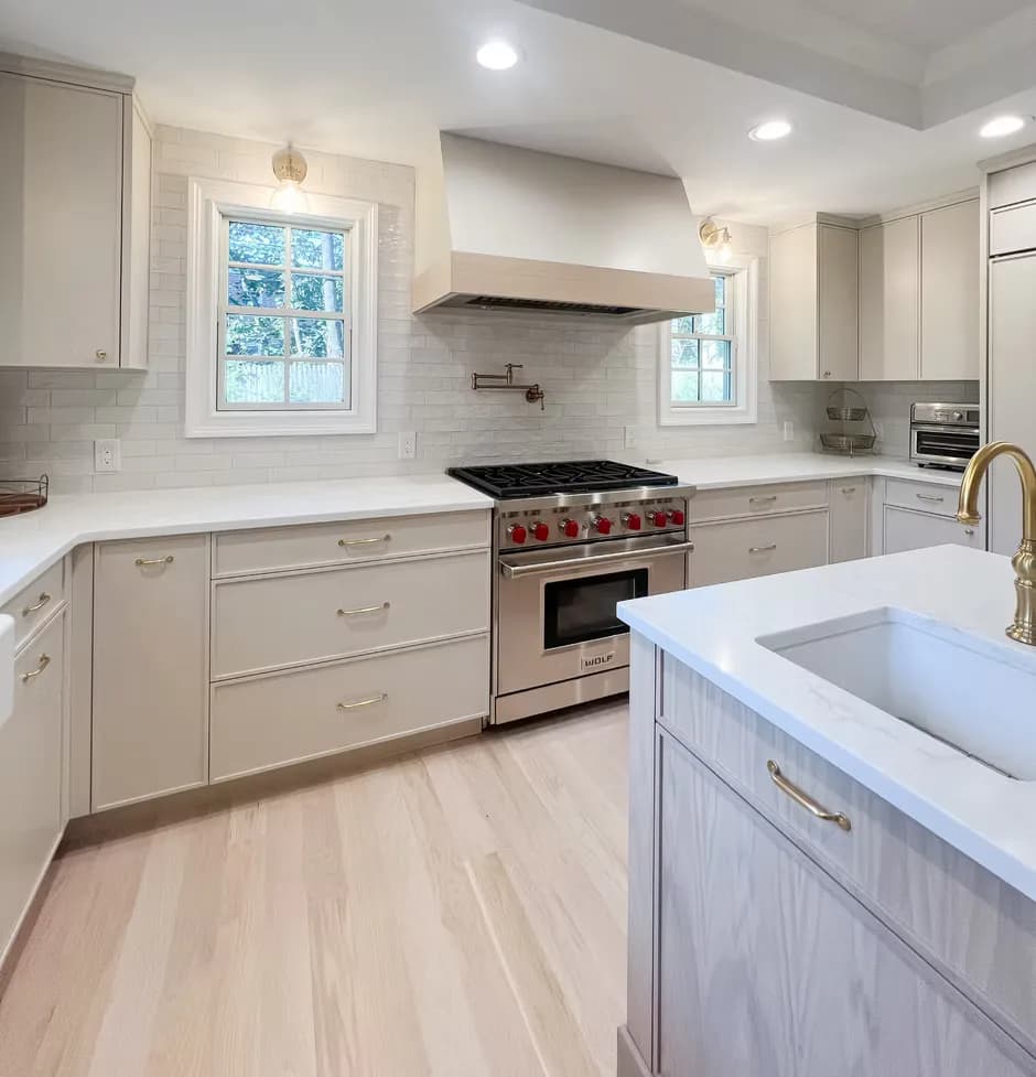 Modern white kitchen with cream cabinetry, white subway tile backsplash, stainless steel range, light wood flooring, and white quartz waterfall island by Melissa McElroy.