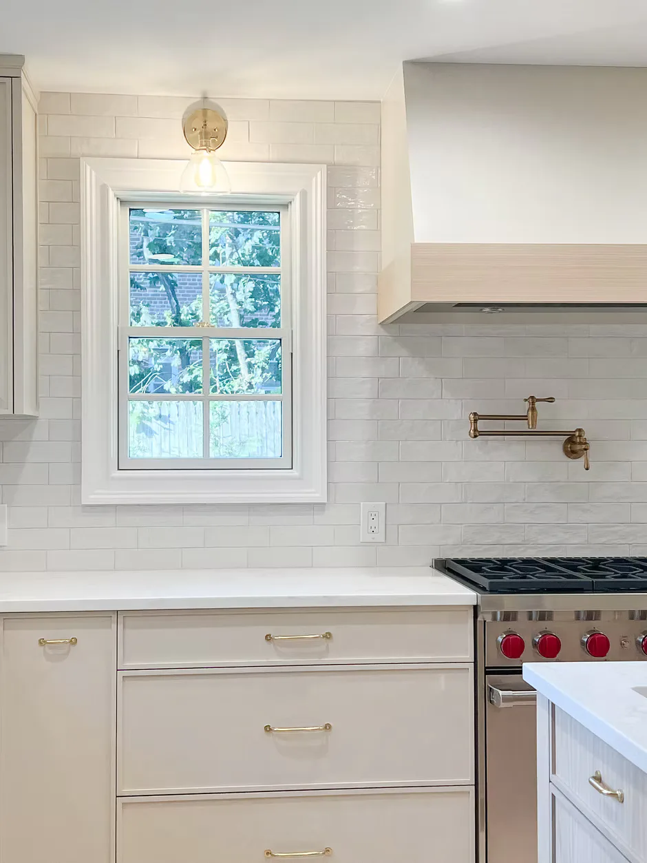 Contemporary kitchen featuring white subway tile backsplash, cream cabinetry with brass hardware, stainless steel range, and large window above sink by Melissa McElroy.