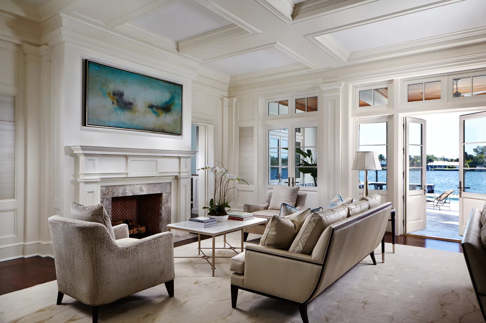 Bright waterfront living room with coffered ceiling, fireplace, neutral furnishings, and extensive glass doors overlooking water views, designed by Melissa Mein.