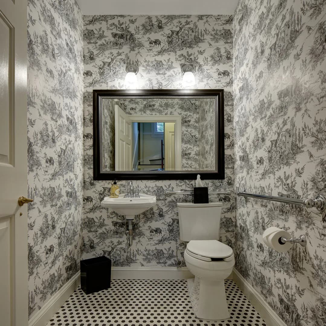 A powder room with black and white toile wallpaper, black-framed mirror, pedestal sink, white toilet, and checkered floor by Melissa Mein.